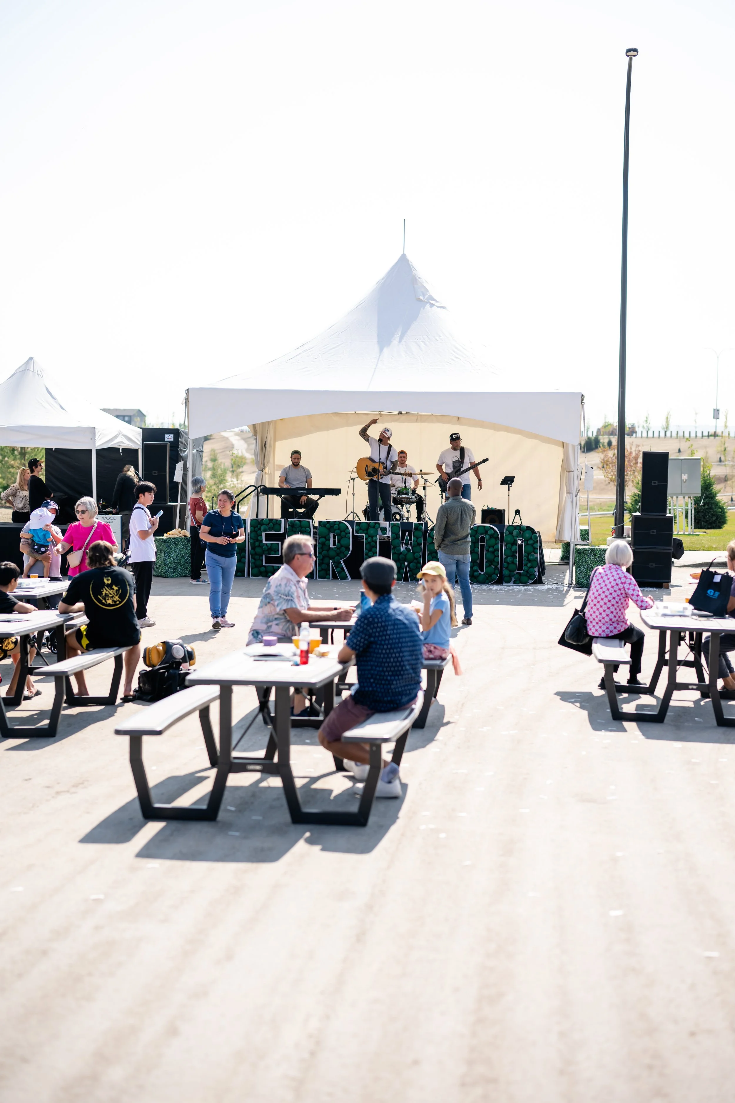 An outdoor concert with a band playing on stage under a white canopy, audience members sitting at picnic tables, and people standing and enjoying the event on a sunny day.
