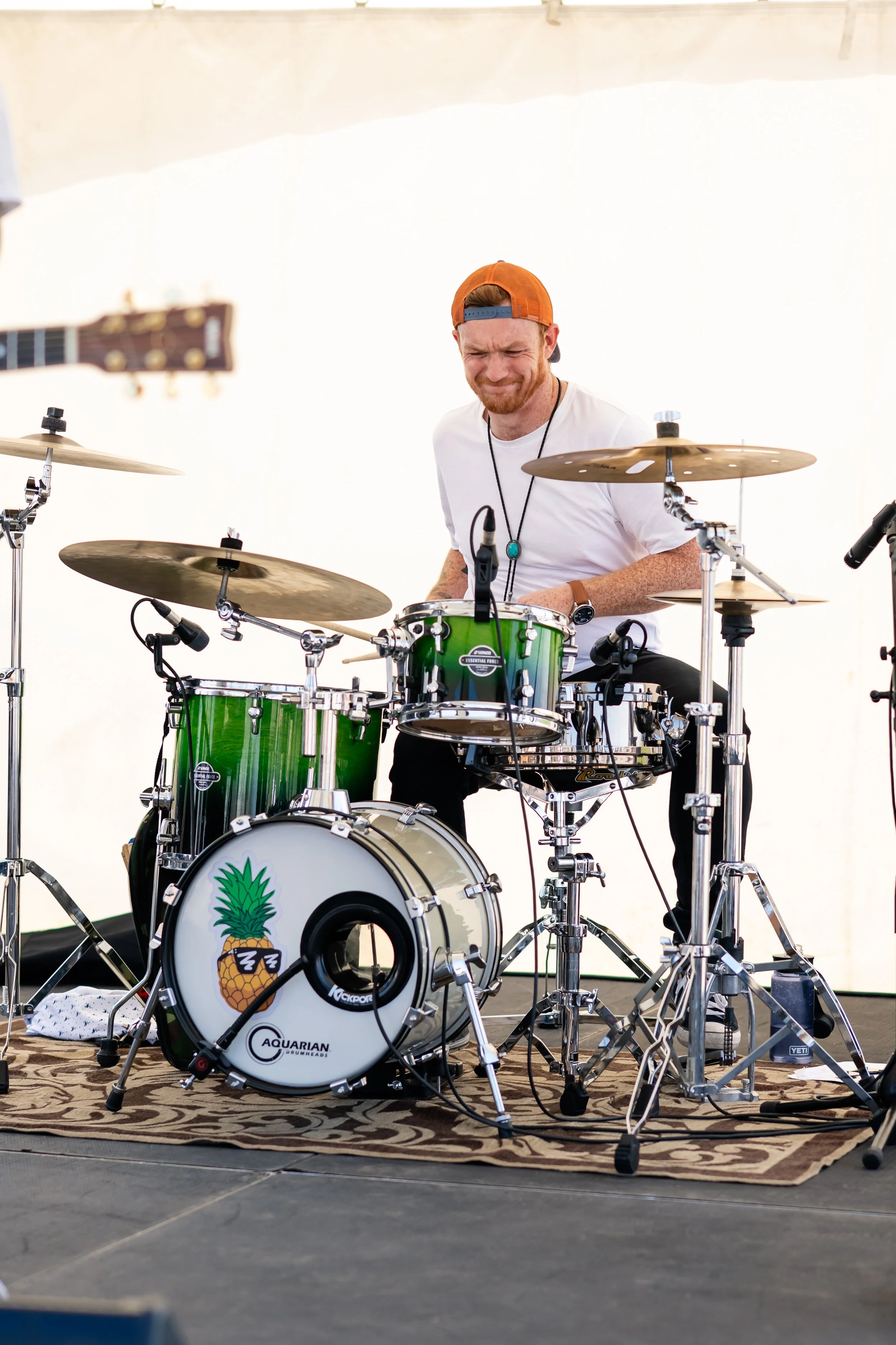 A man with a beard and orange cap playing a green drum set, featuring a pineapple logo on the bass drum, on a stage with a rug and a white background.