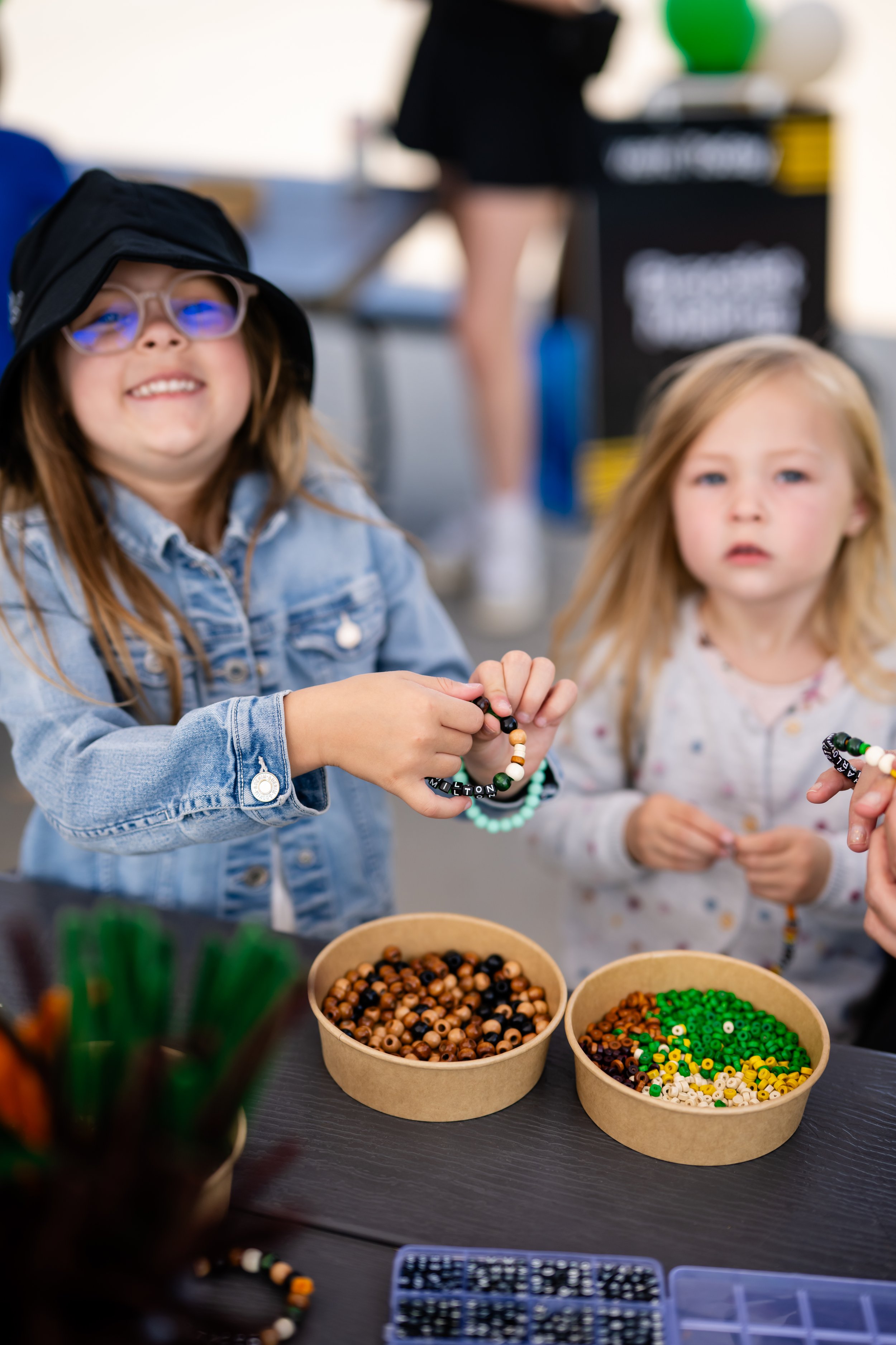 Two young girls with light hair, one wearing glasses and a hat, are making beaded jewelry at a table with bowls of beads in various colors and a bead template on a black table.