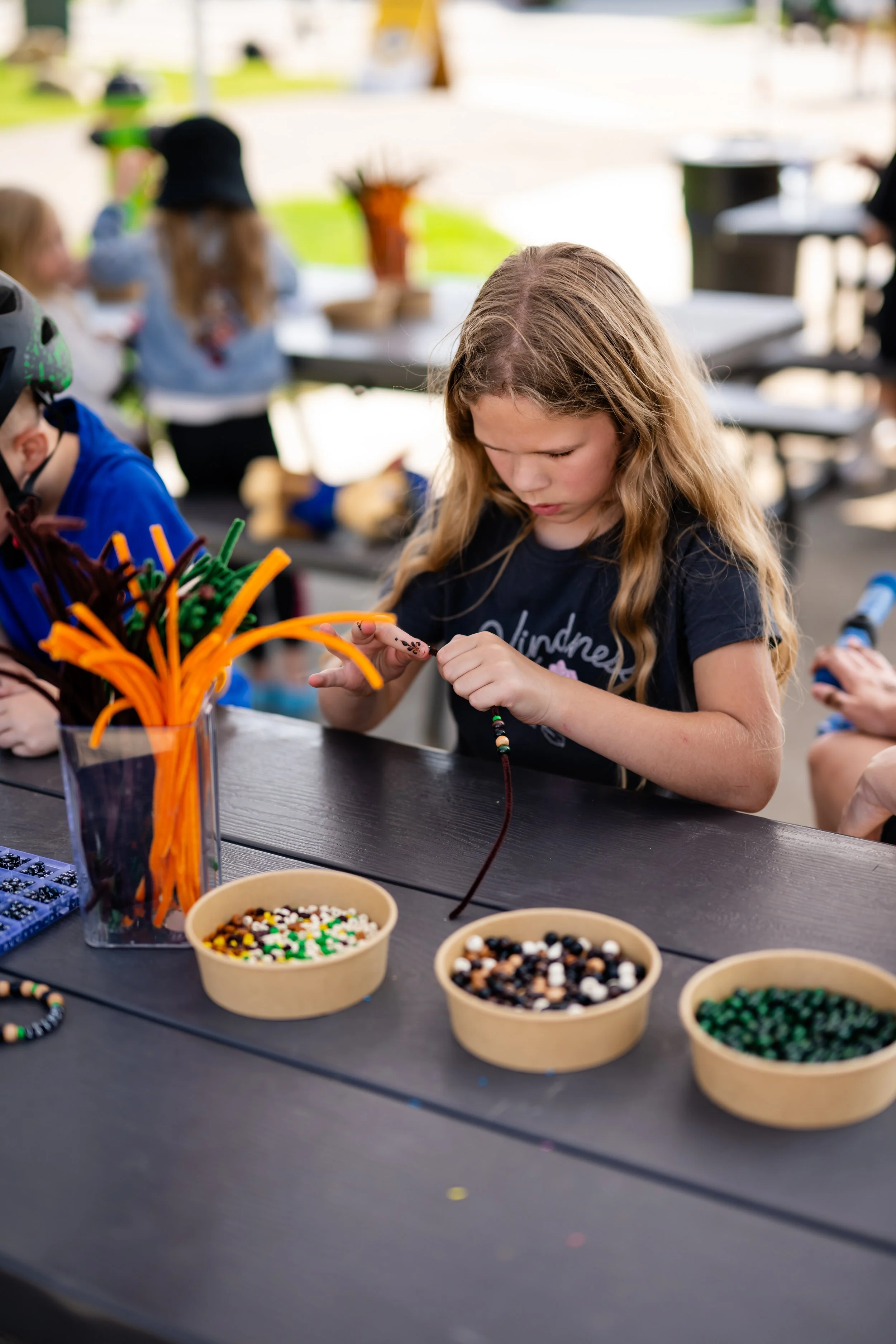 Children sitting at a table outdoors, engaged in arts and crafts activities with cups of beads, pipe cleaners, and decorative items, under a shaded area.