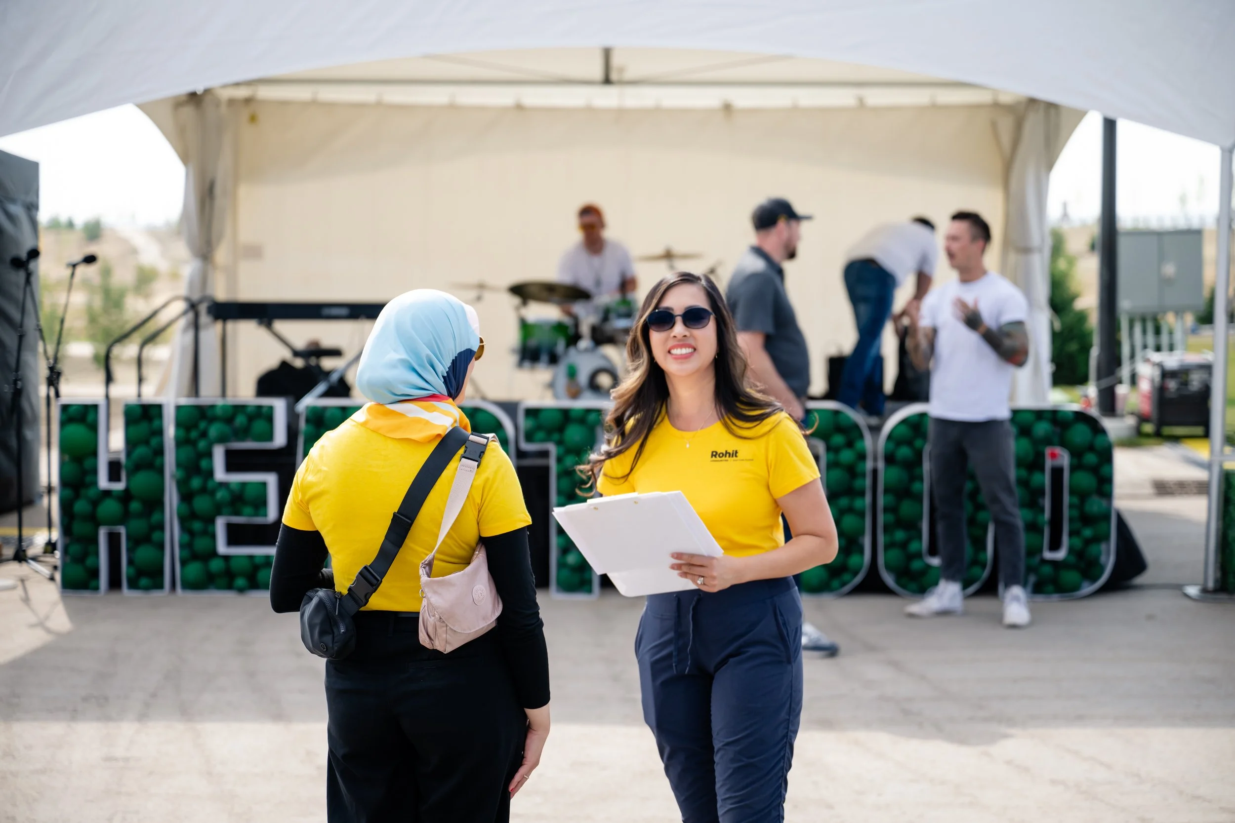 Two women wearing yellow shirts stand and walk outside under a canopy, with a group of men in the background near a green ball toss game, and a musician setup on a stage.