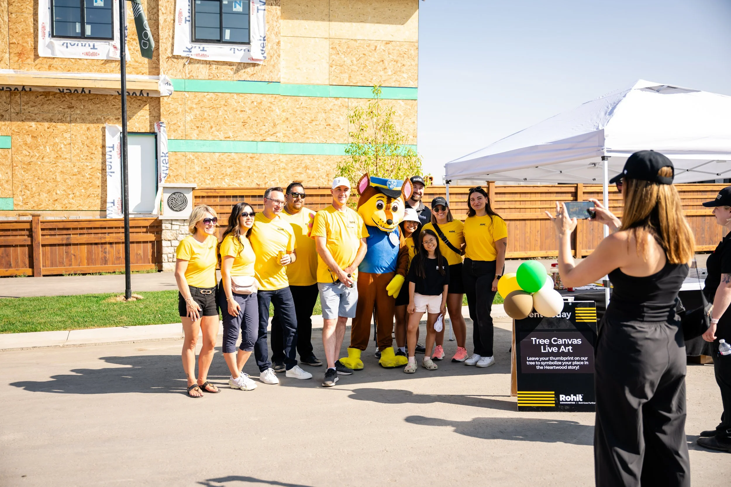 Group of people, including a person in a Paw Patrol mascot costume, standing together for a photo at an outdoor event on a sunny day. Some are wearing yellow shirts and sunglasses, and a woman is taking a picture. There is a white tent and a sign abo