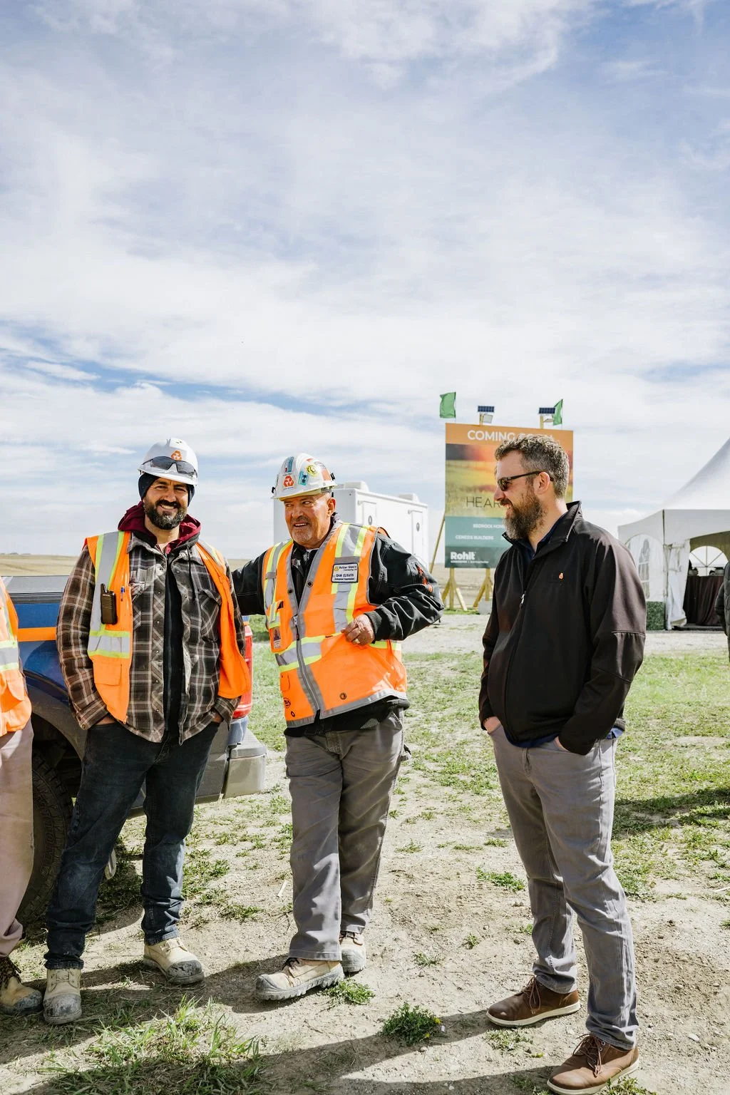 Three men in a construction site; two wearing hard hats and orange safety vests, one in casual clothing. They are standing on dirt near construction tents and signage, under a cloudy sky.