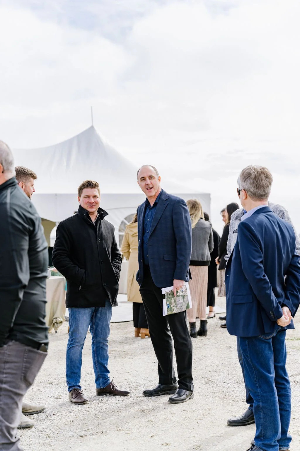 A group of people standing outdoors near a white canopy tent, engaged in conversation. The weather is overcast. Some individuals are wearing coats and jackets, indicating cool weather.