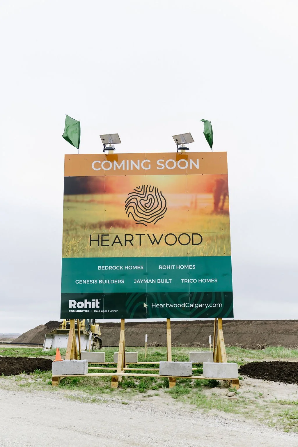 Billboard advertising "Heartwood" community, listing Bedrock Homes, Rohit Homes, Genesis Builders, Jayman Built, Trico Homes. Background of landscape, "Coming Soon" text, and website HeartwoodCalgary.com. Located on a construction site, with equipmen