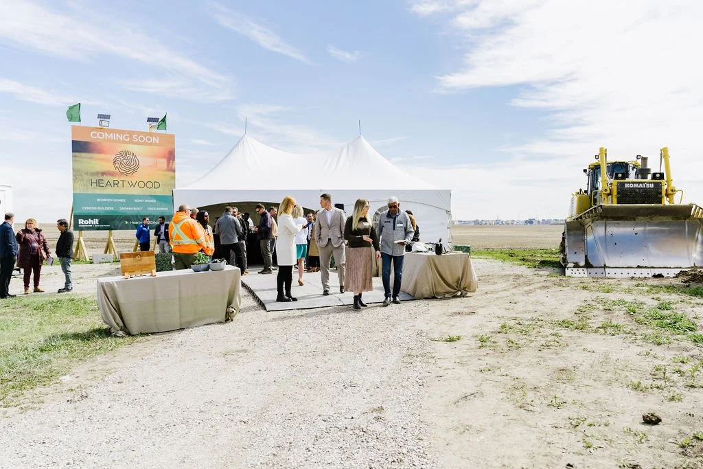 Outdoor event with a white tent, people gathered, and a large construction sign for "Heartwood." Includes a bulldozer and tables with tablecloths.