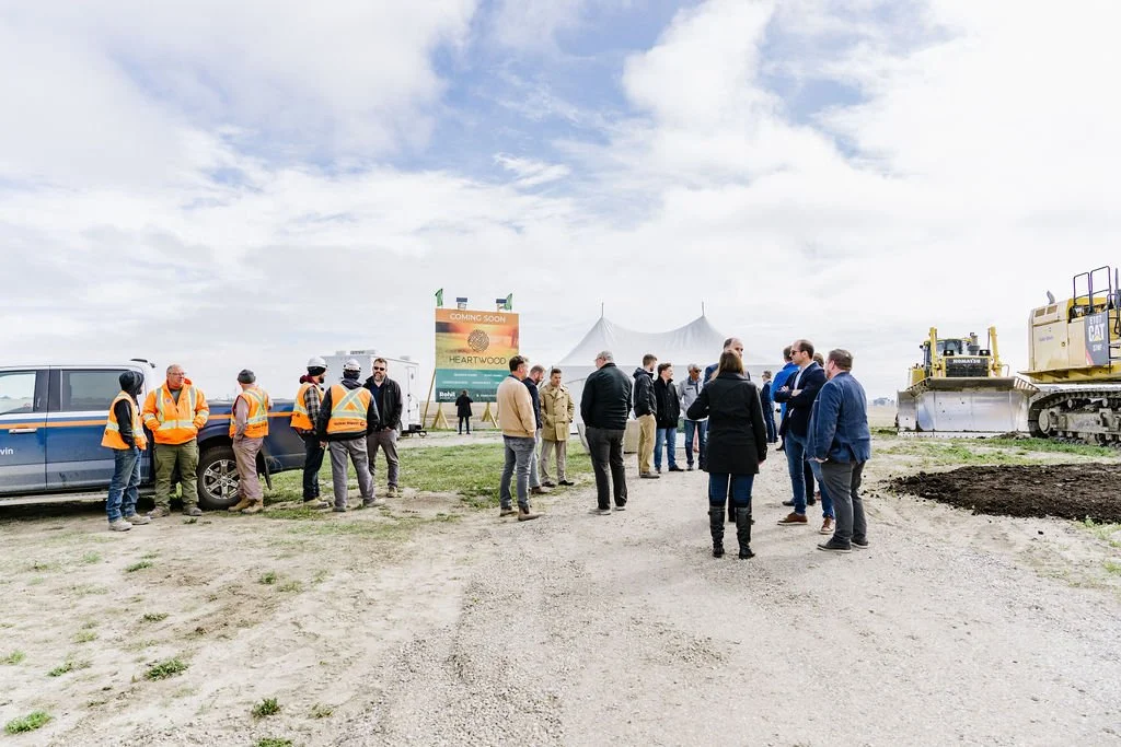 A group of people at a construction site, some wearing high-visibility vests. They are gathered near a sign that reads 'Coming Soon Heartwood' in a field with construction equipment and vehicles nearby.
