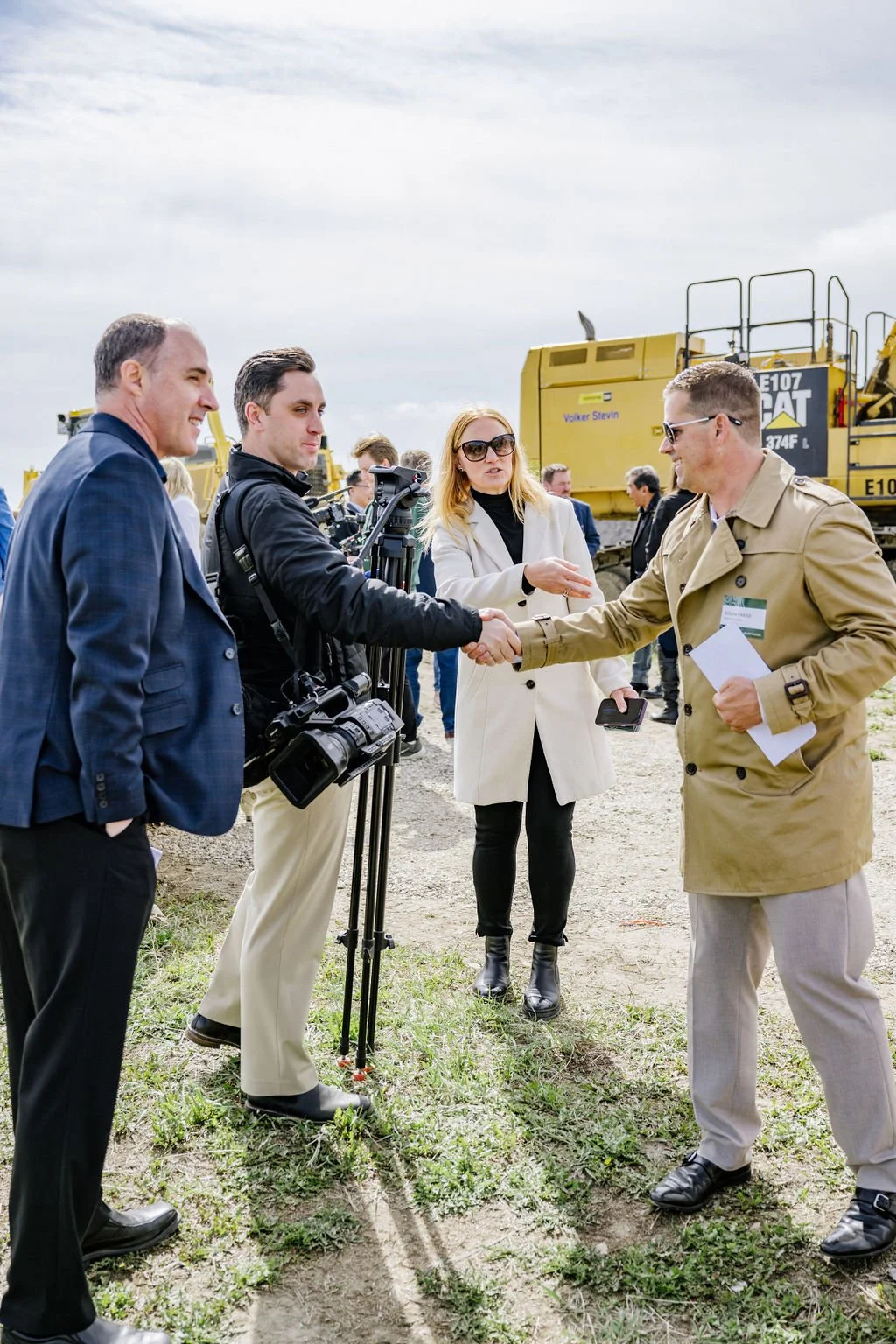 Group of people outdoors, one holding a camera, others shaking hands, construction equipment in the background.