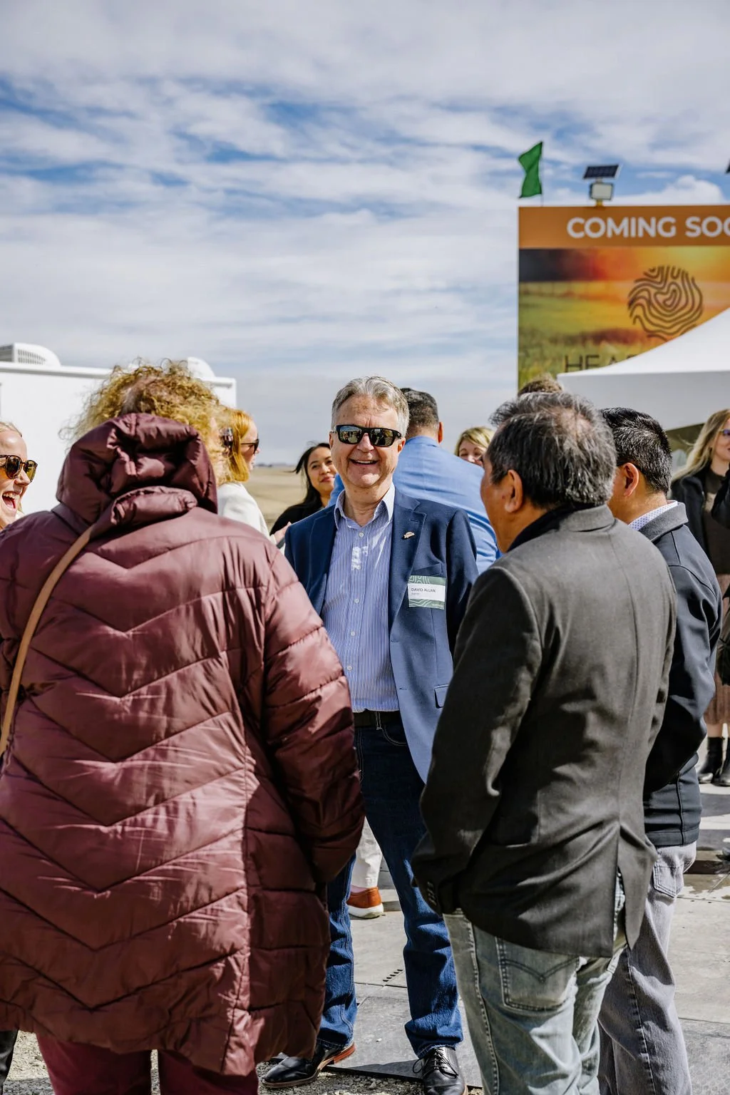A group of people socializing outdoors under a cloudy sky, with one person in a blue blazer and sunglasses smiling. A sign in the background says "Coming Soon."