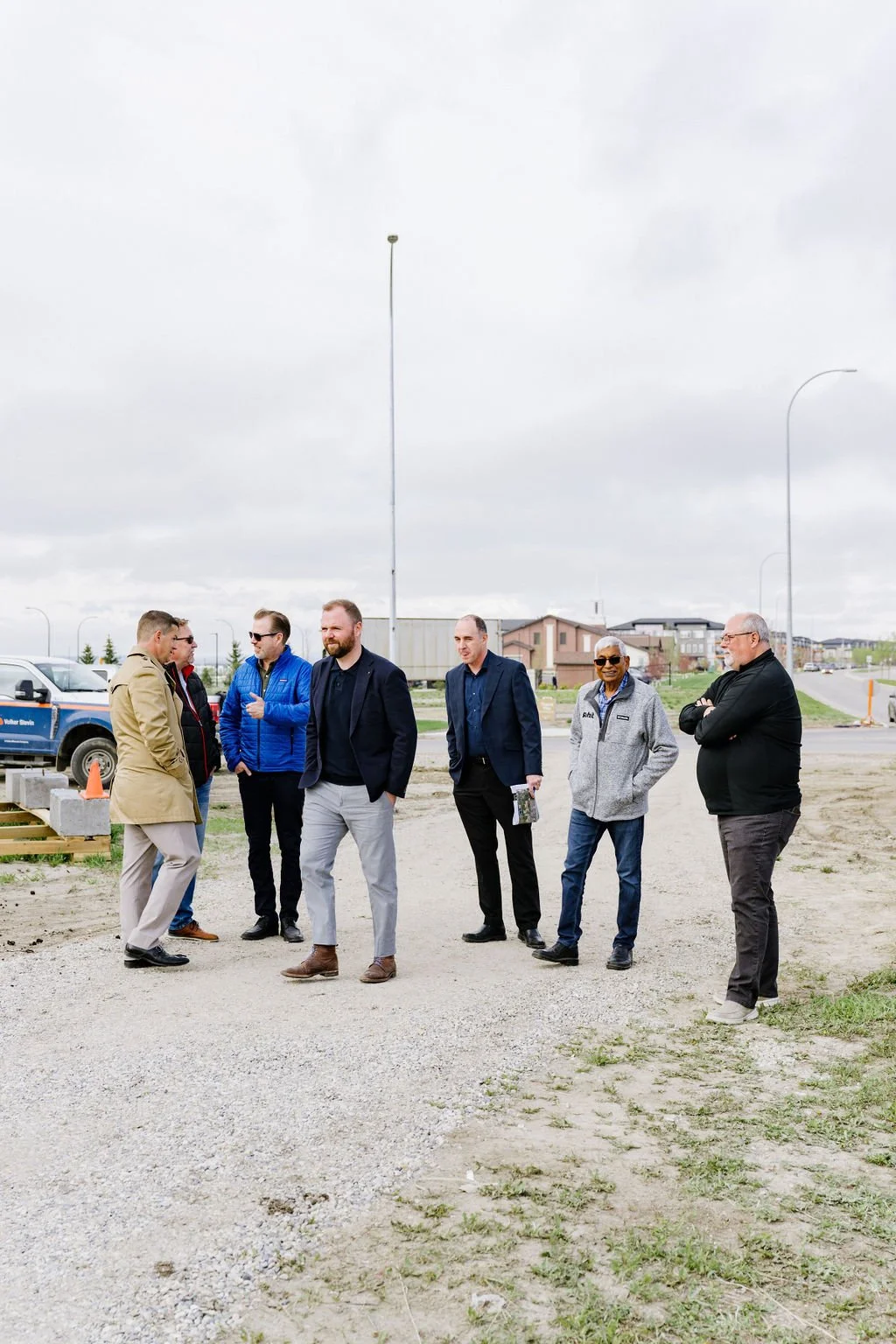 Group of men standing on a construction site, overcast sky, wearing casual and formal attire.