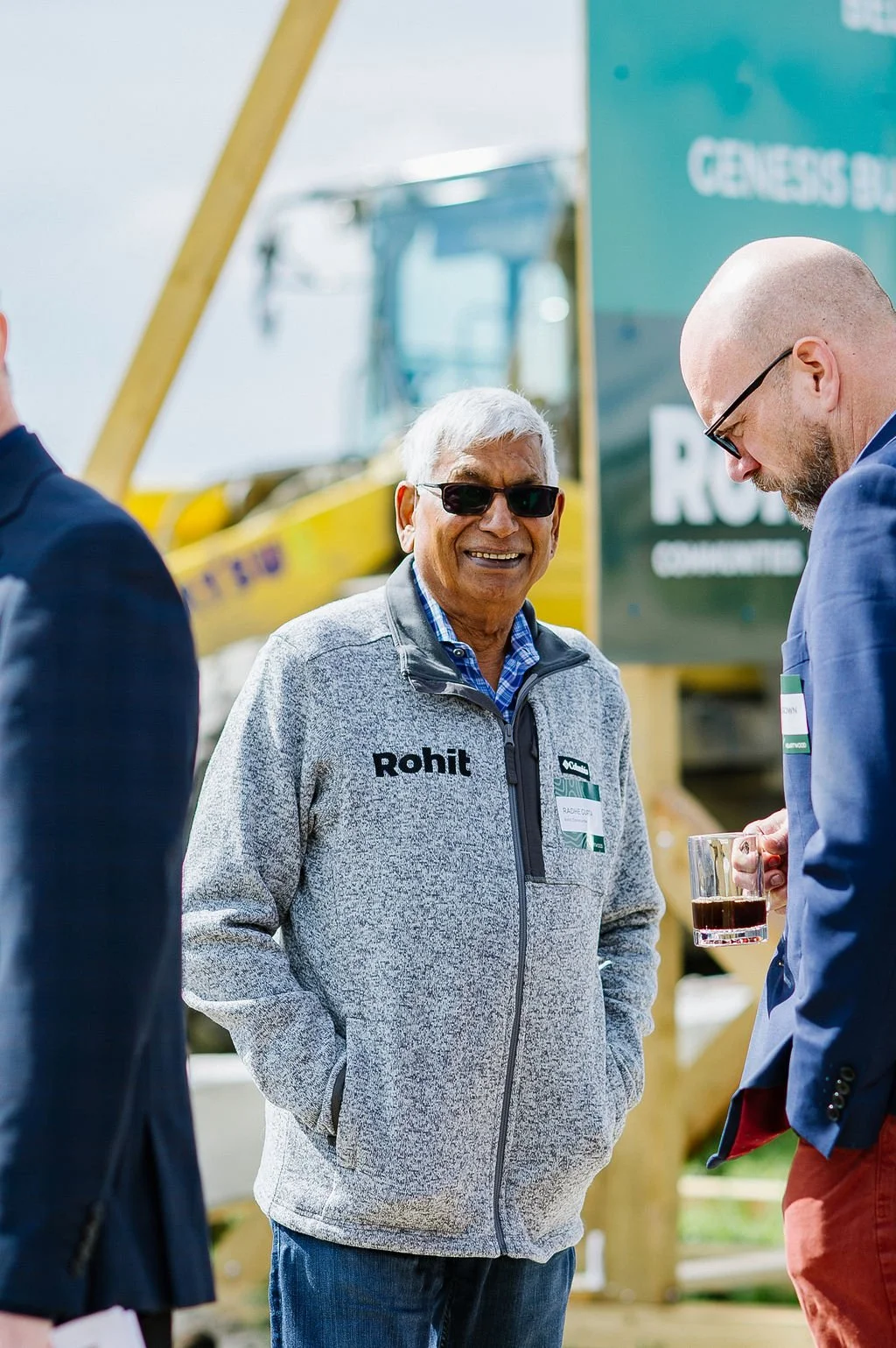Two men in outdoor setting, one wearing sunglasses and a gray jacket with "Rohit" logo, smiling, the other holding a glass. Construction equipment and a sign in the background.