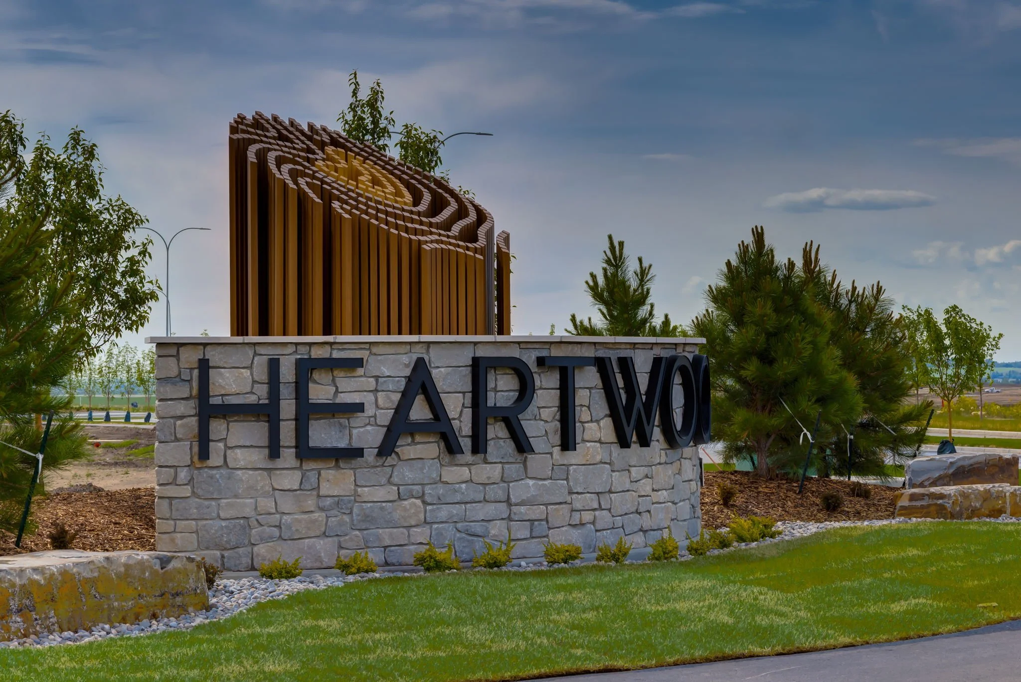 Sign that reads 'HEARWOOD' with a decorative wooden logo on a stone base, surrounded by trees and landscaping under a partly cloudy sky.
