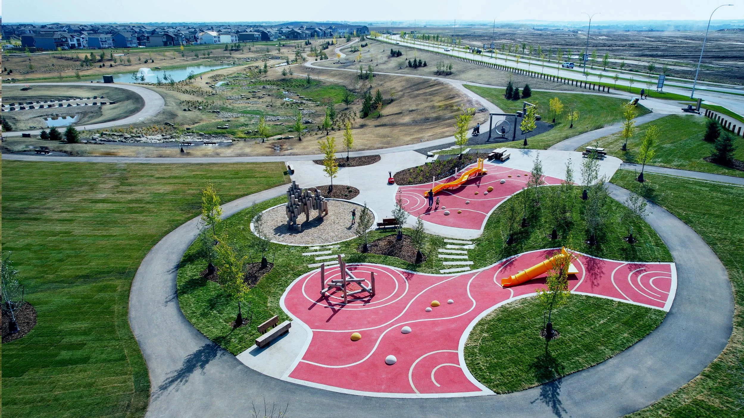 Aerial view of a modern park with walking paths, young trees, playground equipment, a pond, a lawn, and a slide, with residential houses and a highway in the background.
