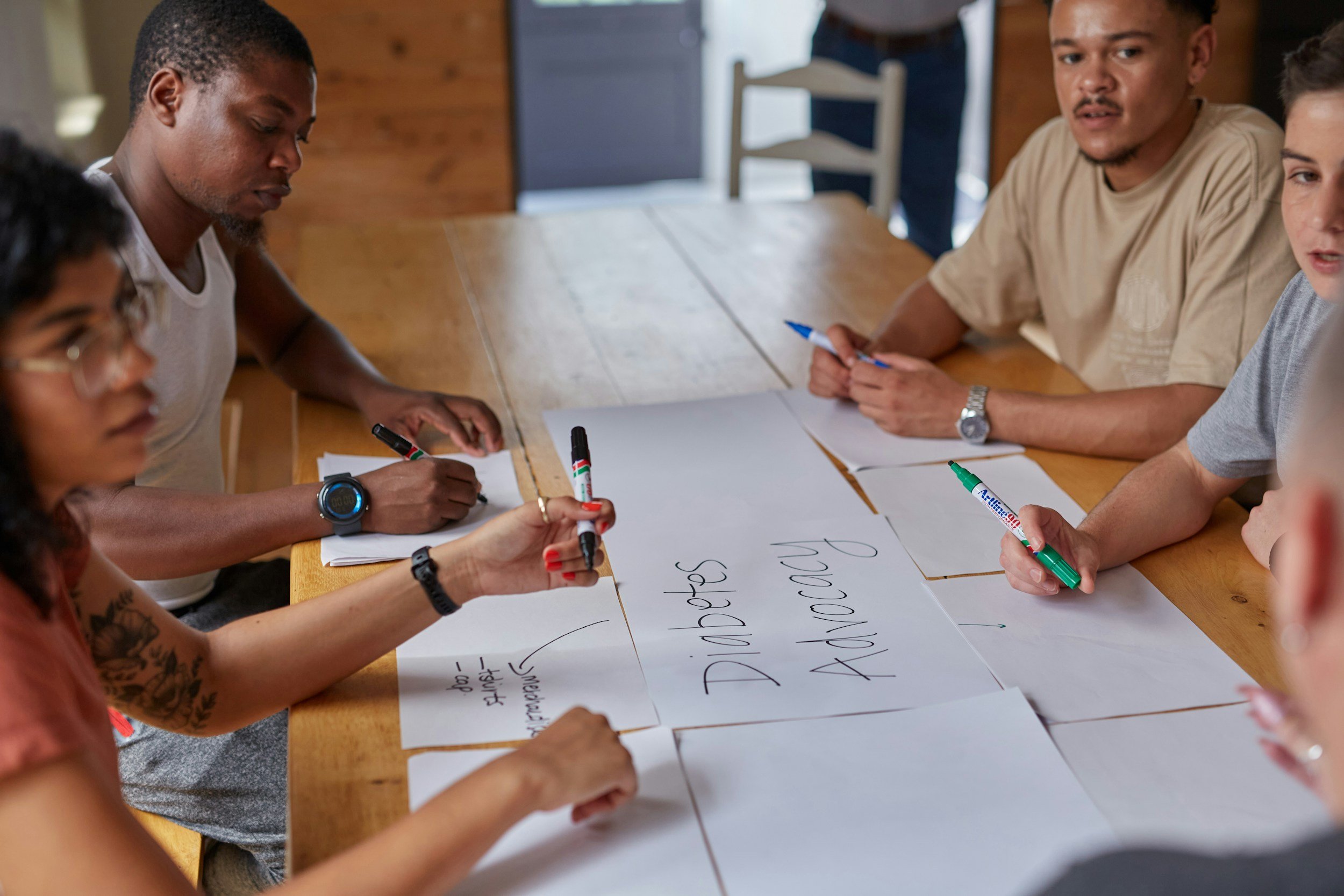 A diverse group of people sitting around a wooden table participating in a collaborative activity, with some writing on paper and large sheets of paper with handwritten notes on the table, in a cozy, rustic room.
