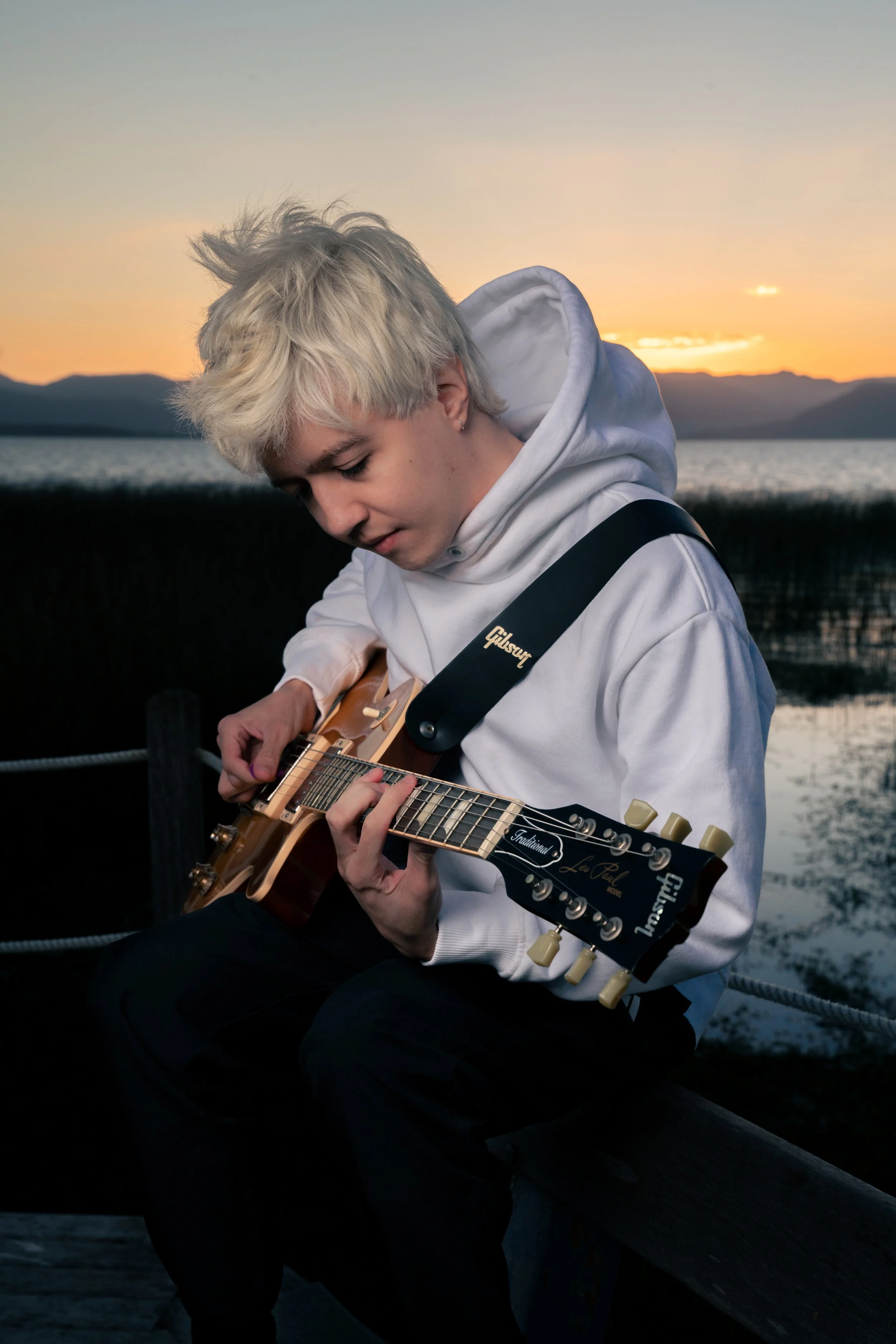 Jovem tocando violão ao pôr do sol, vista para um lago e montanhas ao fundo.