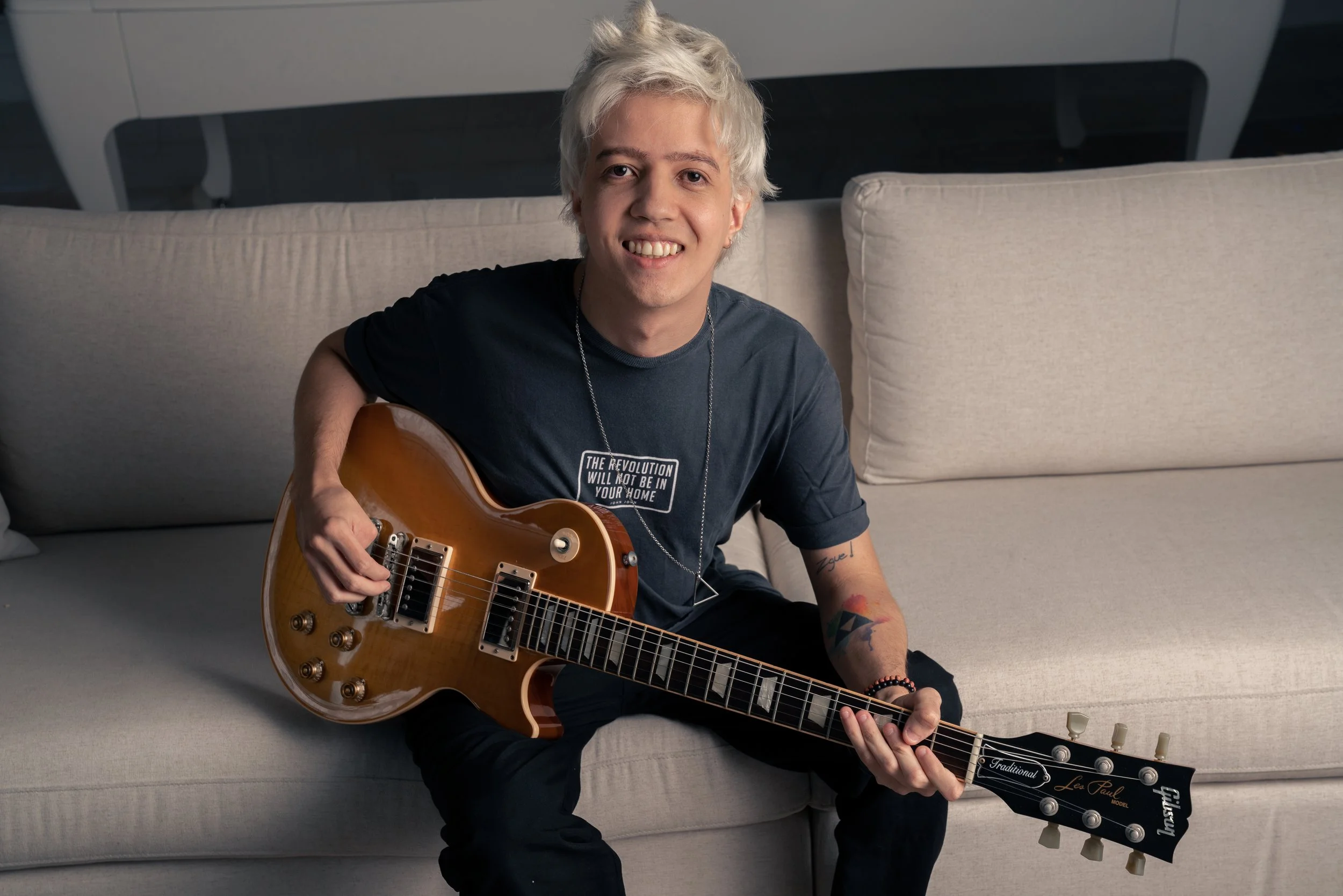 Homem com cabelo branco e roupas pretas sentado no sofá, segurando uma guitarra elétrica de madeira e sorrindo para a câmera.
