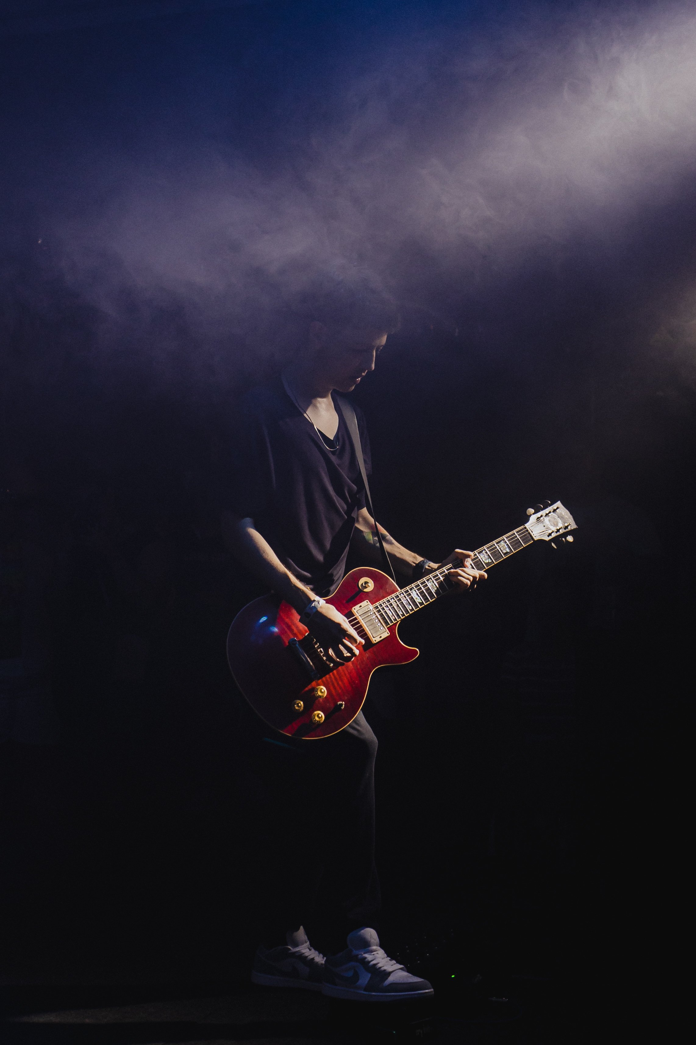 Pessoa tocando guitarra vermelha em um palco escuro, usando tênis brancos e calça escura.