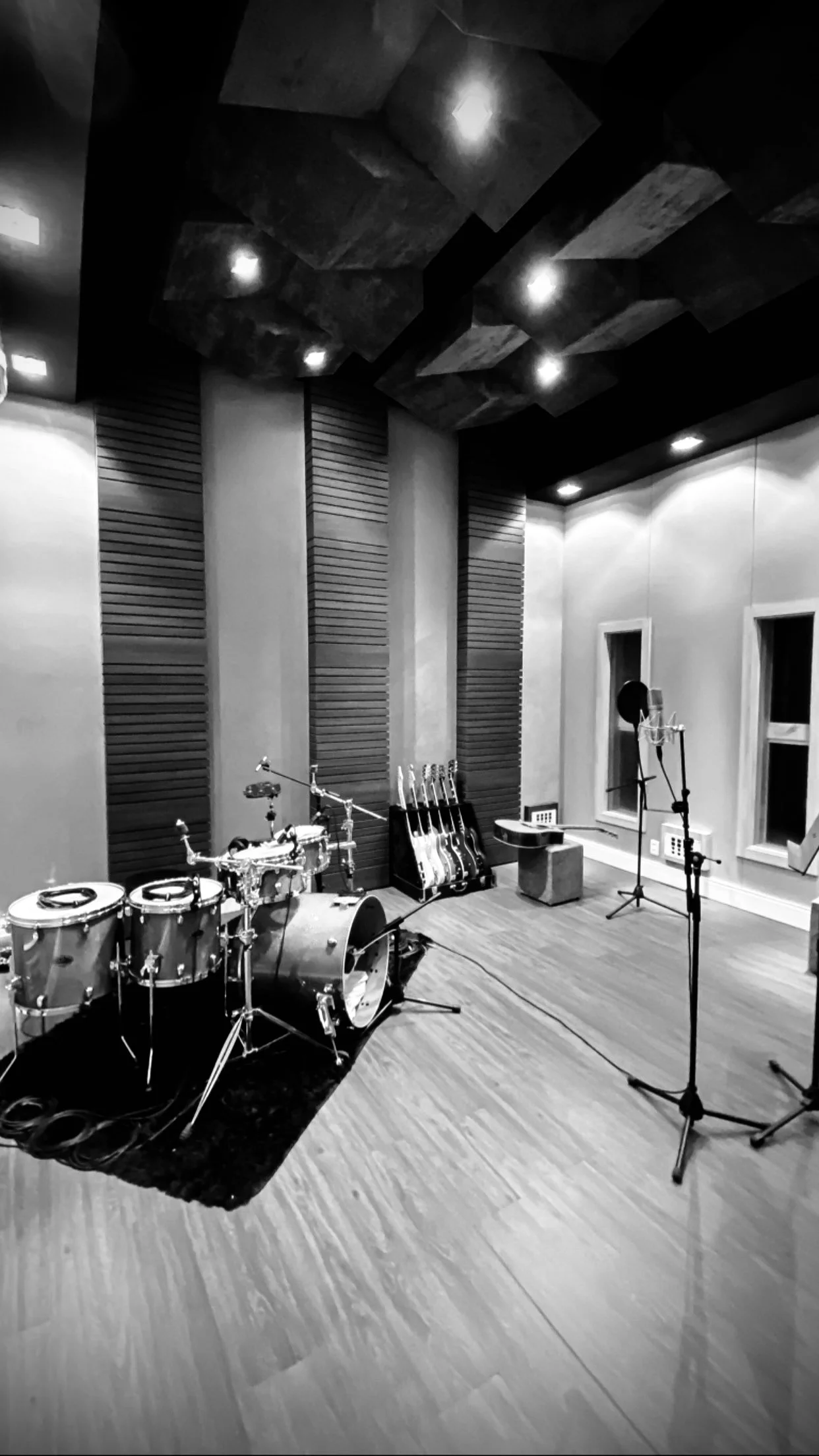 Sala de gravação de estúdio musical com instrumentos musicais, microfones e equipamentos de som, paredes acústicas de madeira e teto com iluminação embutida.