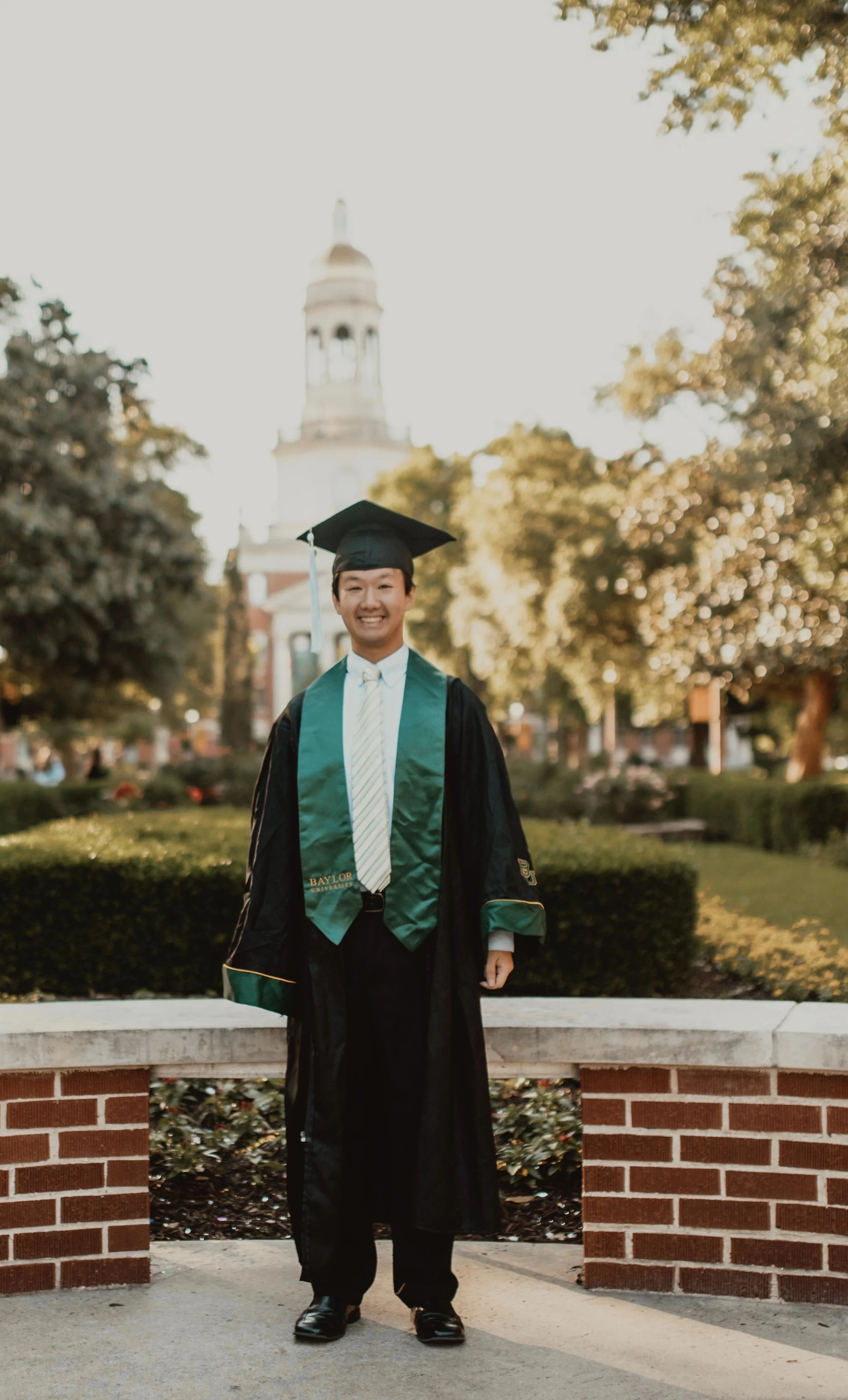 A young man in graduation attire, including a cap and gown, standing outdoors in front of a brick wall. There are trees and a building with a clock tower in the background, bathed in warm sunlight.
