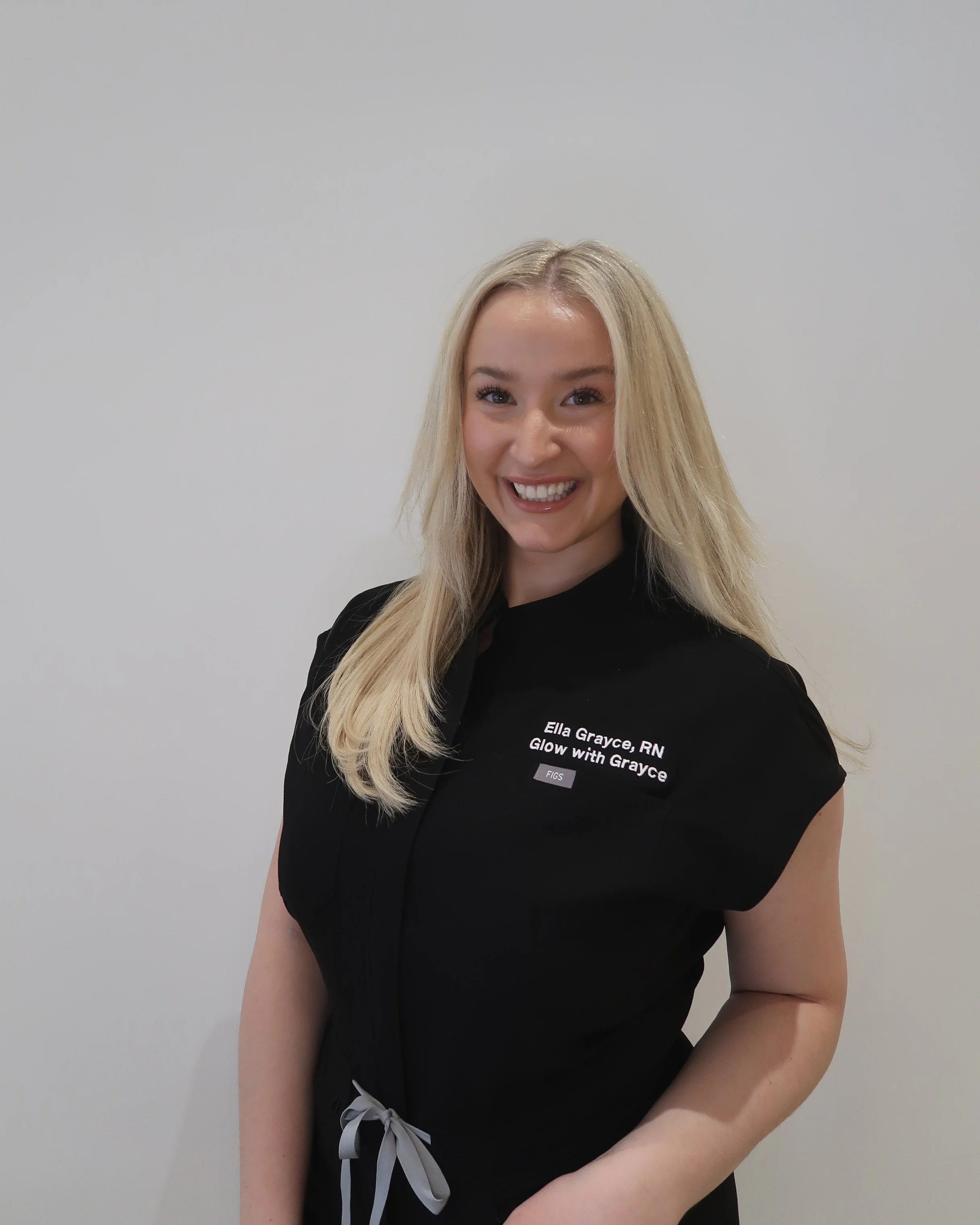A smiling woman with blonde hair wearing a black shirt with text, standing against a plain white wall.