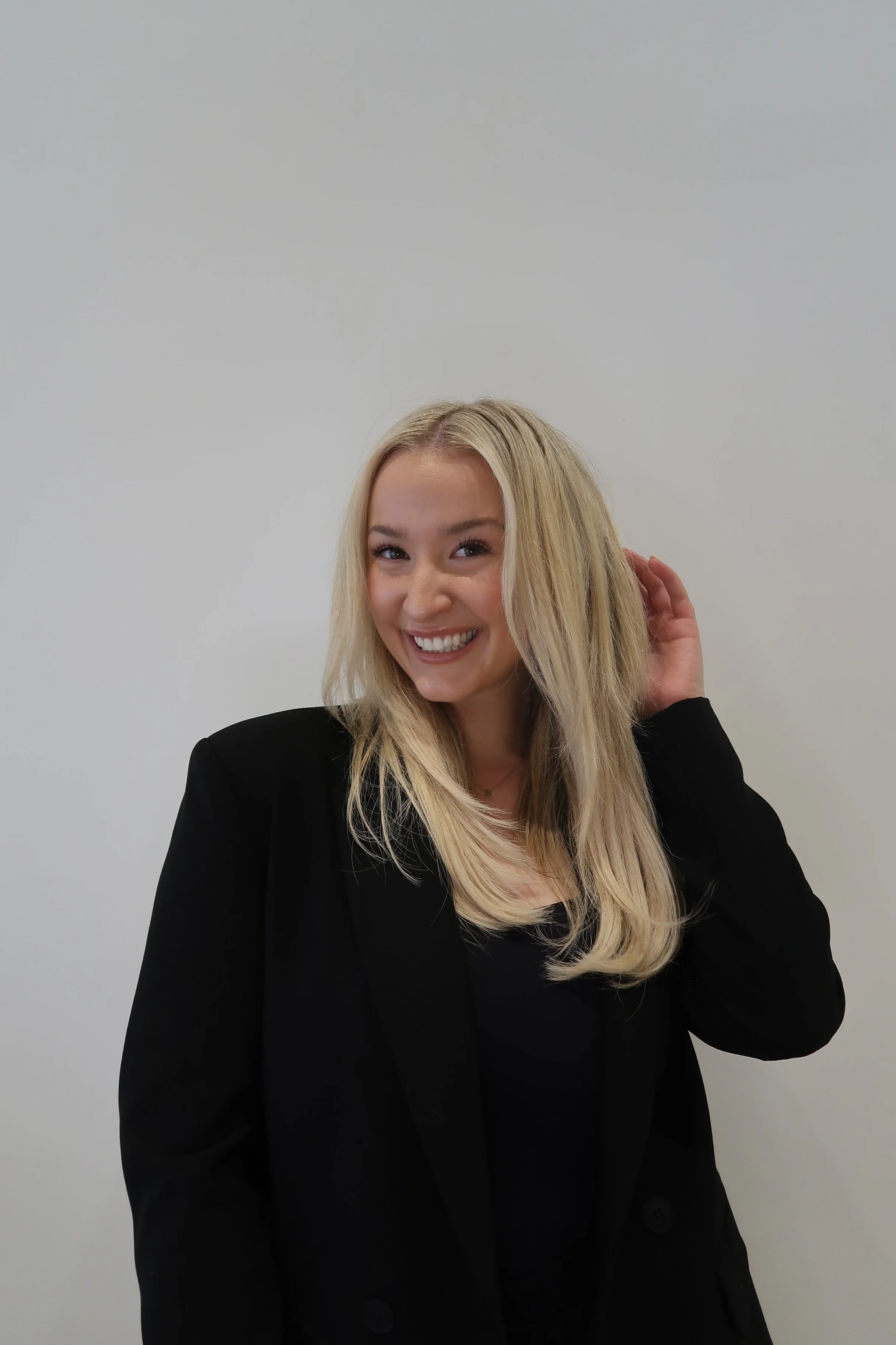 A woman with long blonde hair, wearing a black blazer, smiling, and adjusting her hair against a plain light gray background.
