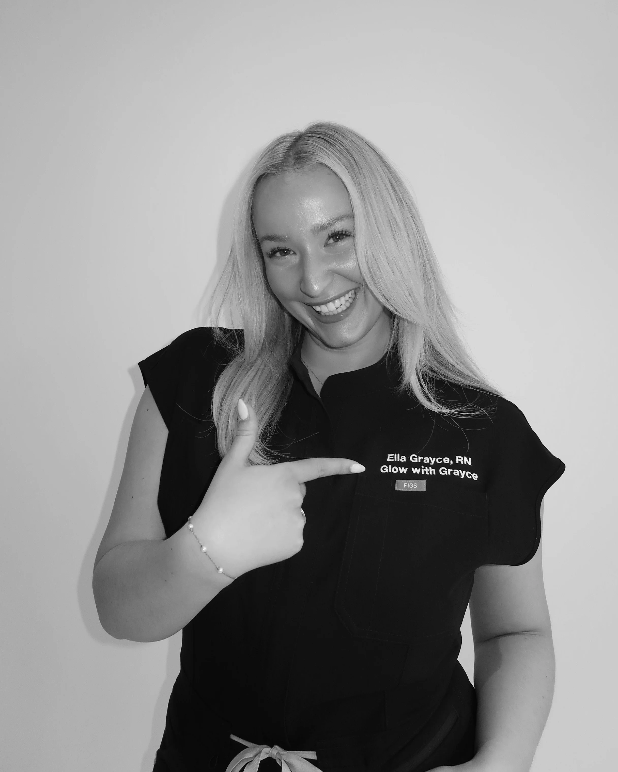 A smiling woman with long blonde hair, wearing a dark shirt with a name tag that says 'Ella Grayce, RN Glow with Grayce,' making a finger gun gesture against a plain background.