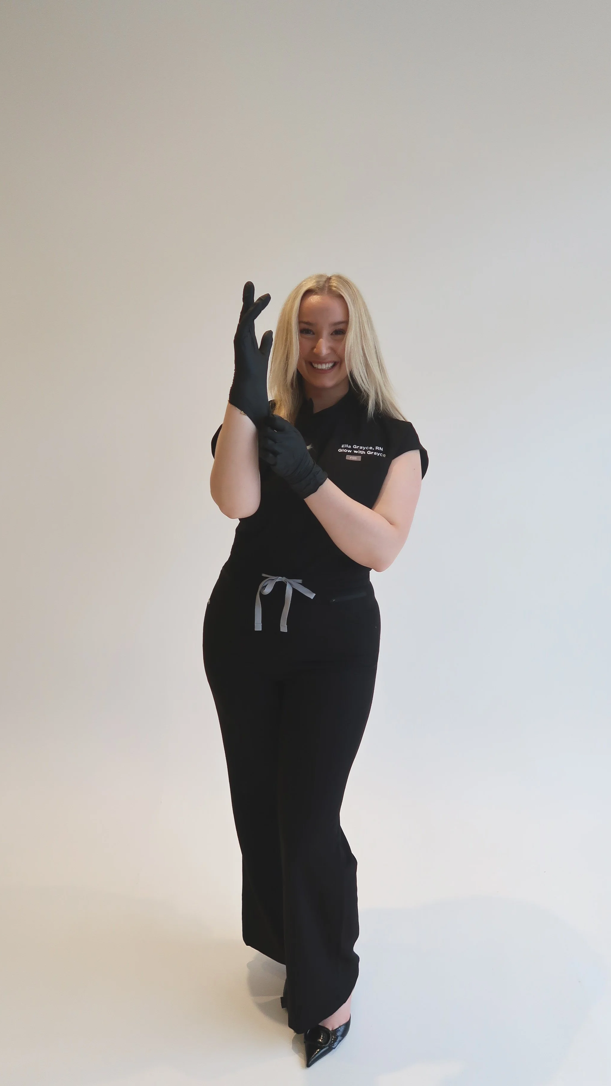 A smiling blonde woman dressed in black with black gloves, standing against a plain light-colored background.