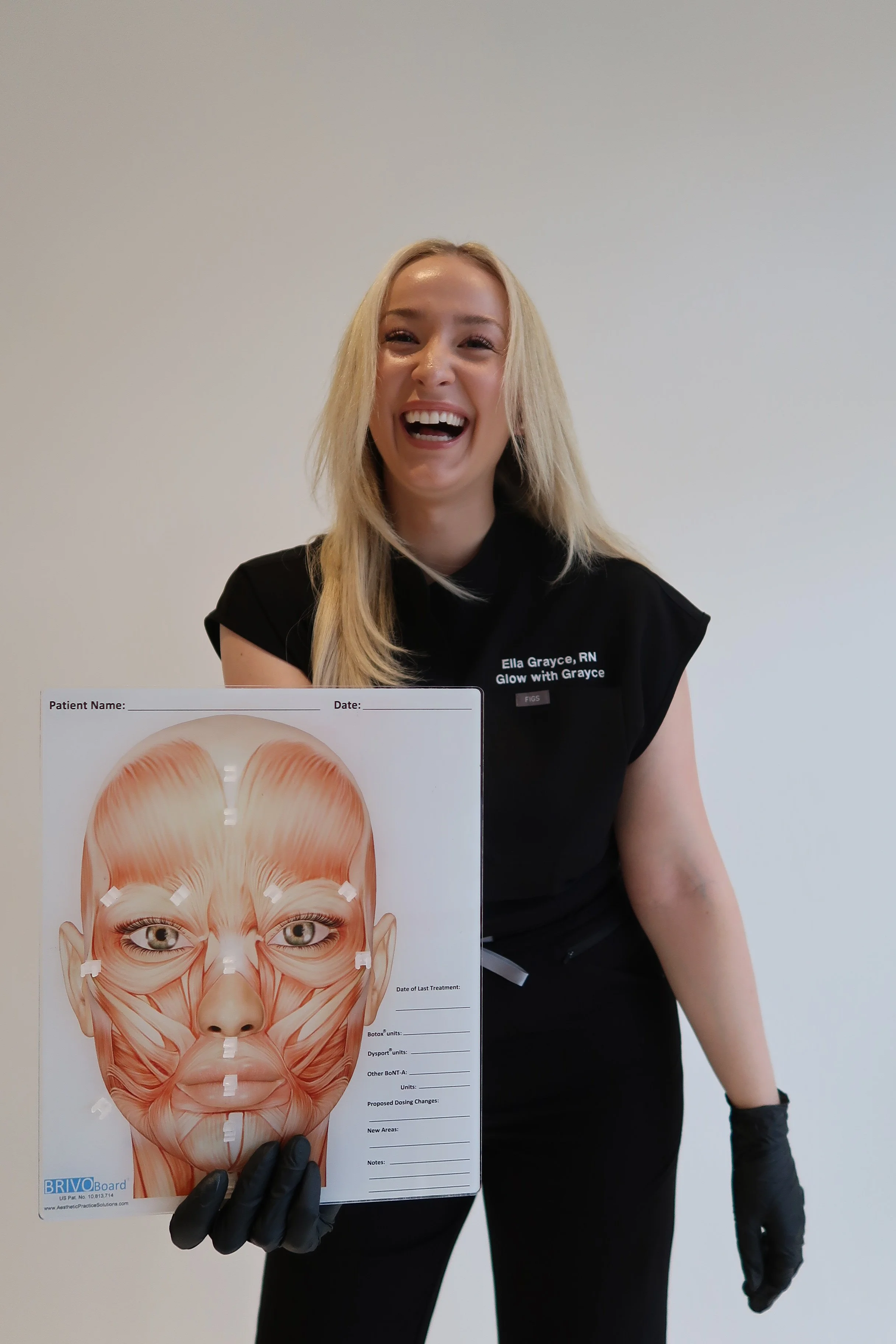 A woman holding a facial anatomy chart in front of a plain wall, smiling and wearing black gloves and a black shirt with her name and credentials. The chart shows detailed facial muscles and anatomy.