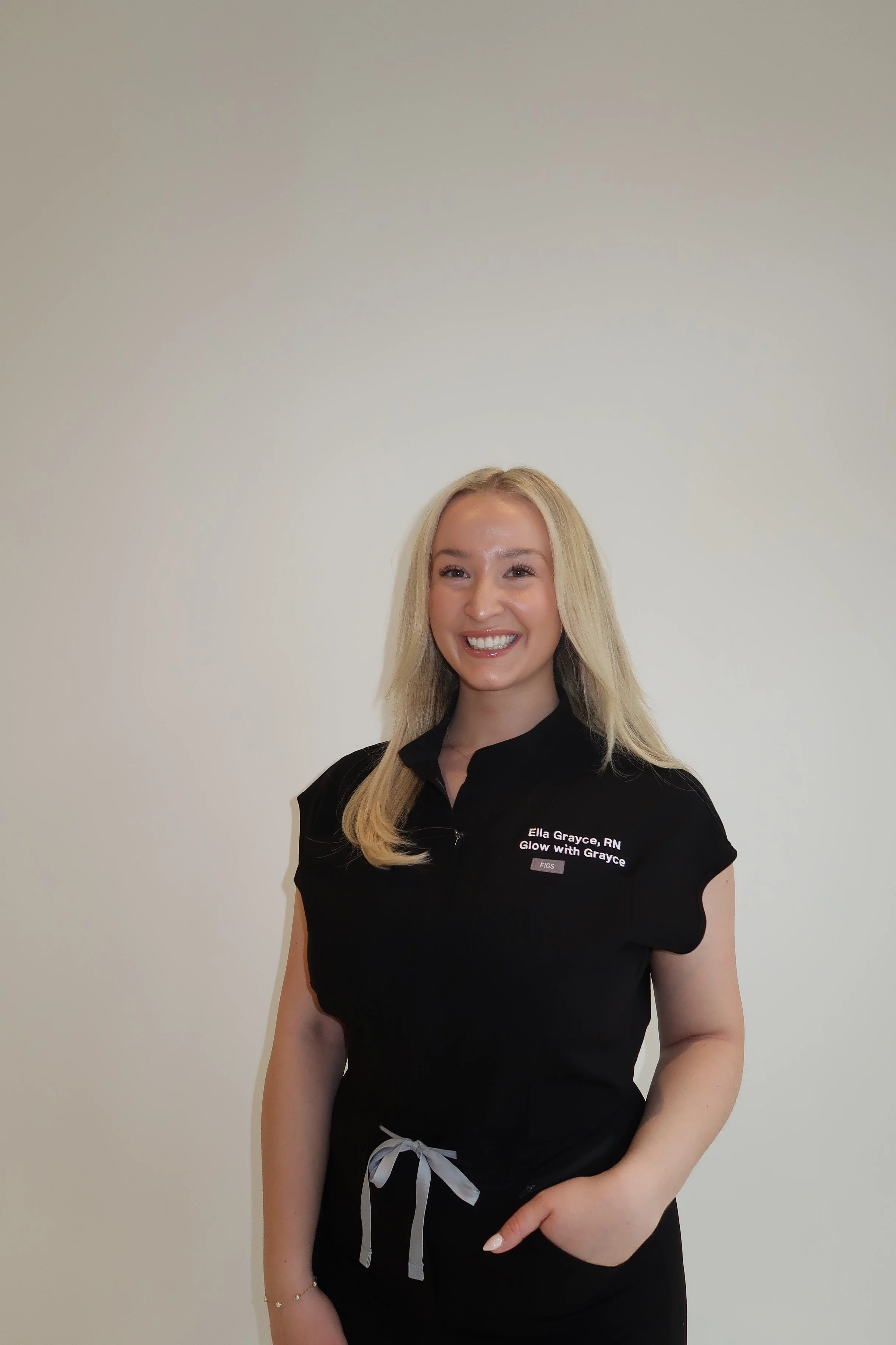 A smiling woman with long blonde hair wearing a black top with a name tag that reads "Ella Grayce, RN, Glow with Grayce" standing against a plain light-colored wall.