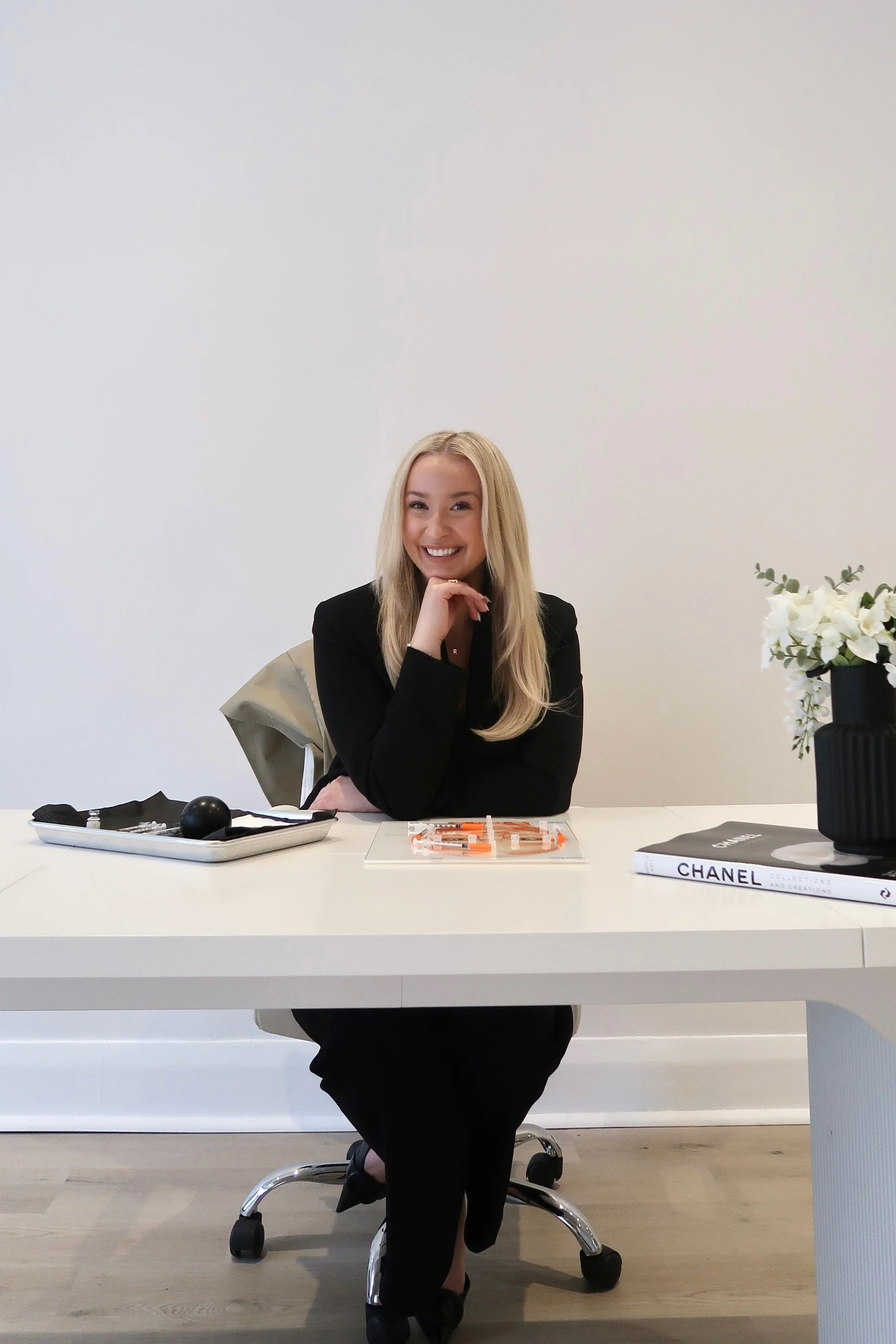 Smiling woman with long blonde hair sitting at a white desk with a black and white book labeled 'CHANEL', a black bowl, and lip gloss, wearing a black blazer in a modern office with a white wall and a vase of white flowers on her right.