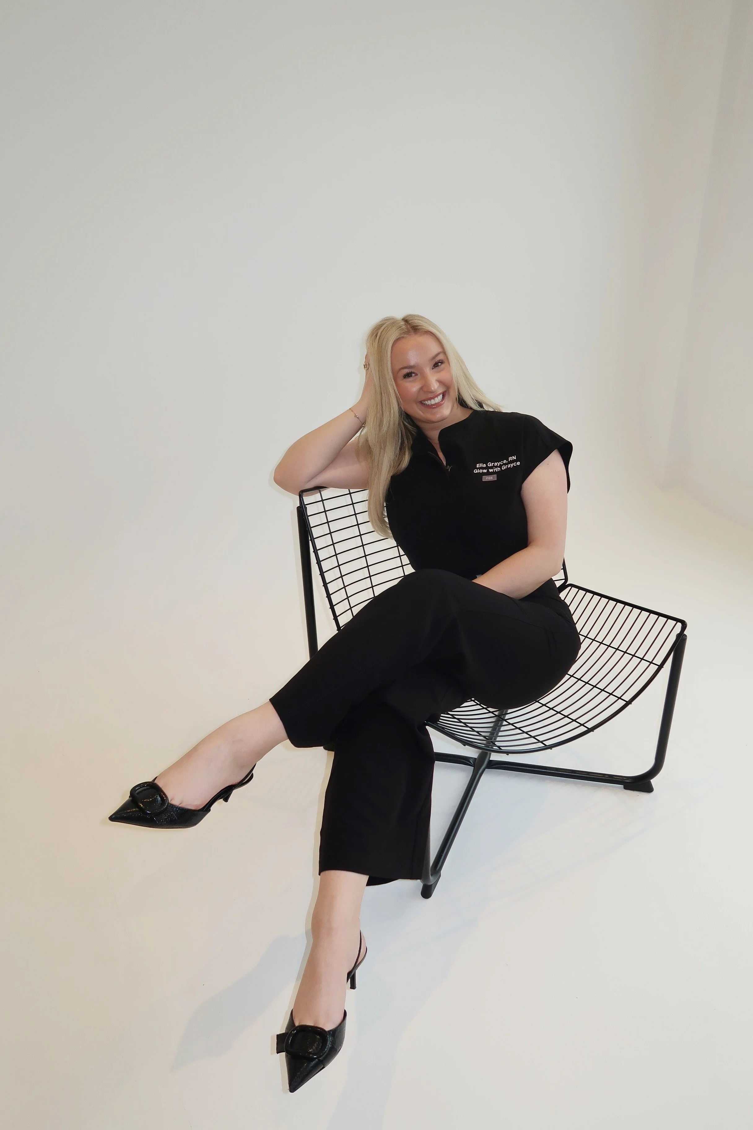 A woman with long blonde hair sitting on a black wire chair against a plain white background, wearing black pants, black high heels, and a black top with white text, smiling and posing with her left arm resting on her head.