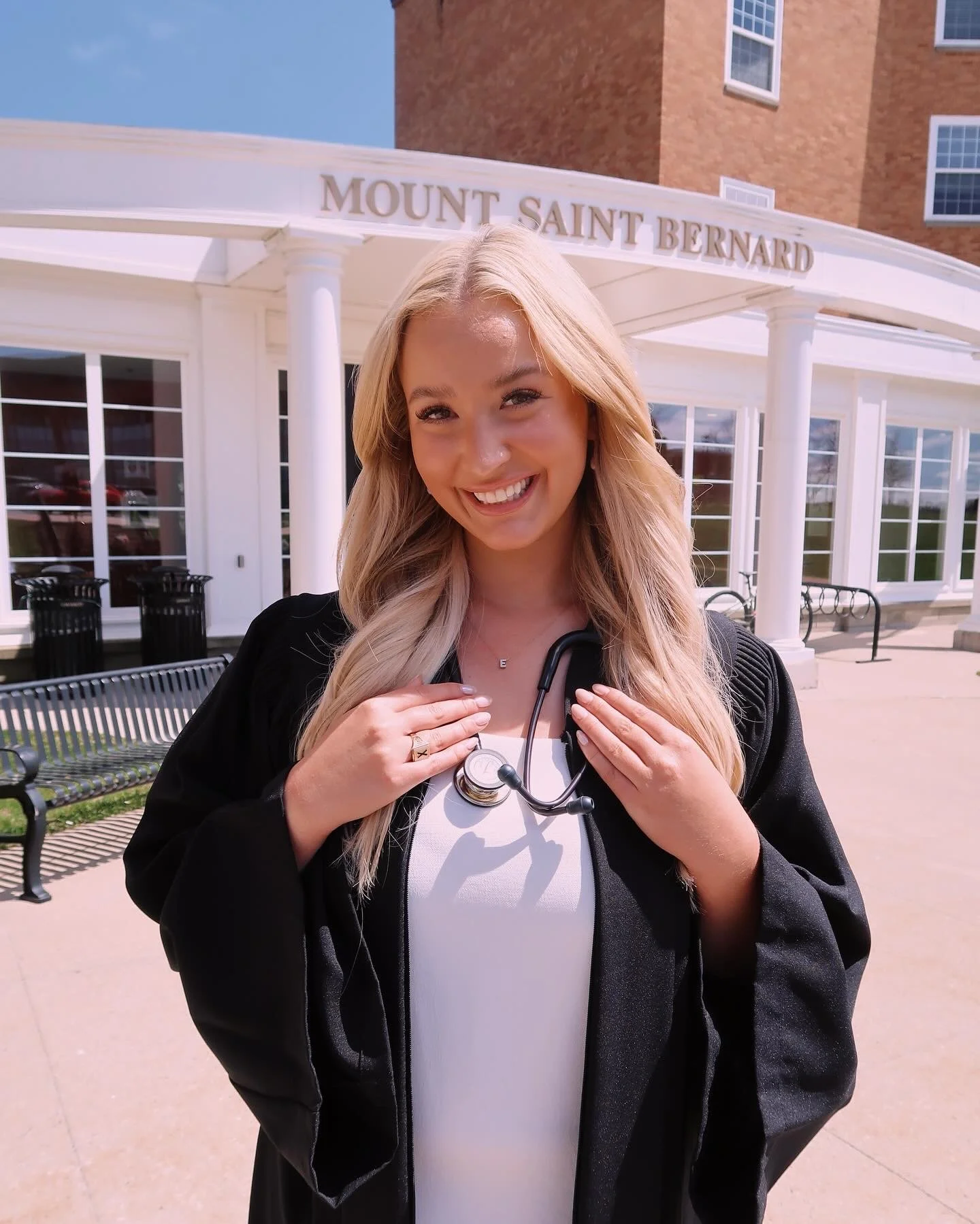 A young woman with long blonde hair smiling outside of a building with a sign that says "Mount Saint Bernard." She is wearing a white top, a black jacket, and has a stethoscope around her neck.