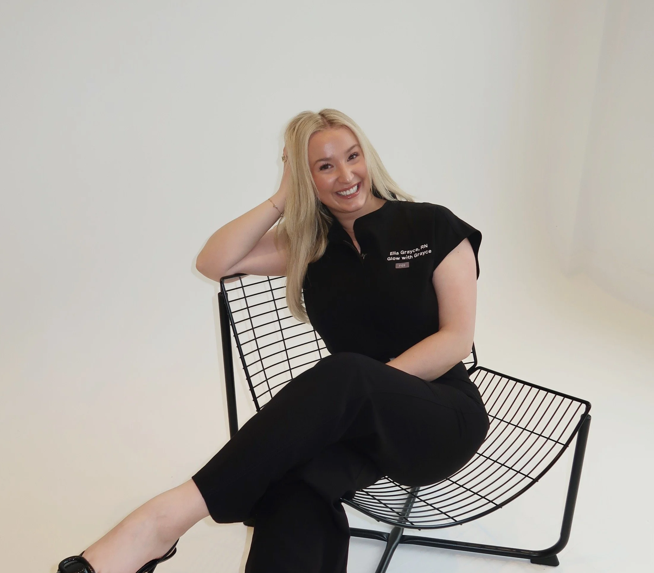 A young woman with long blonde hair sitting on a black wire chair, smiling and touching her hair, against a plain white wall background. She wears a black shirt with text and black pants.