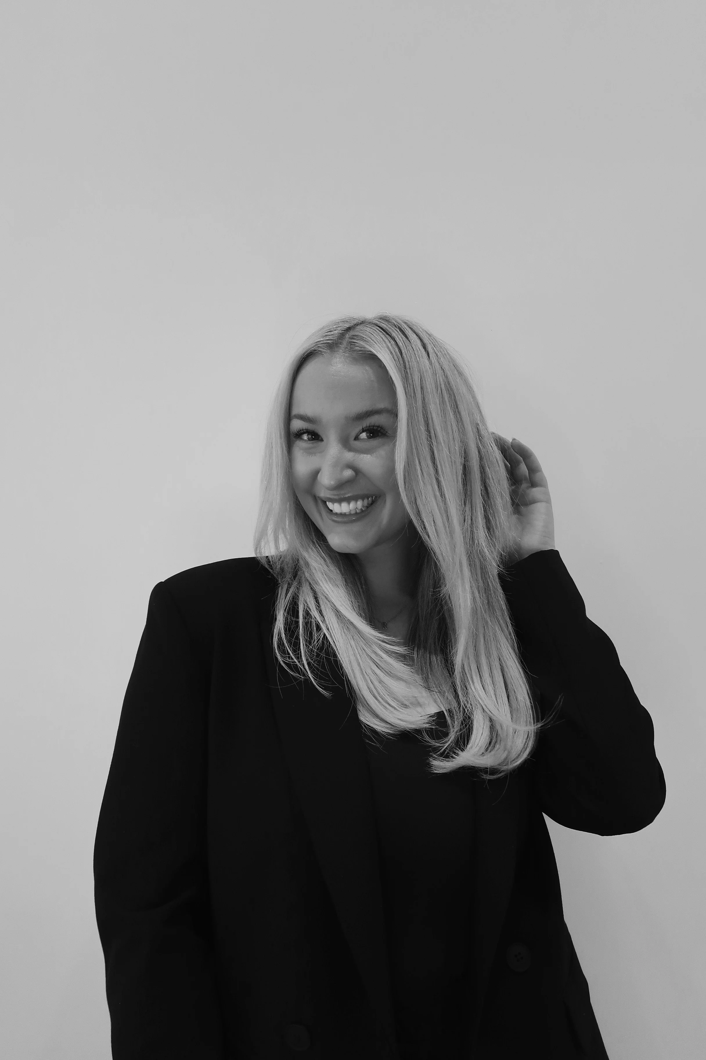 Black and white photo of a woman with long hair smiling and touching her hair, wearing a dark blazer against a plain wall.