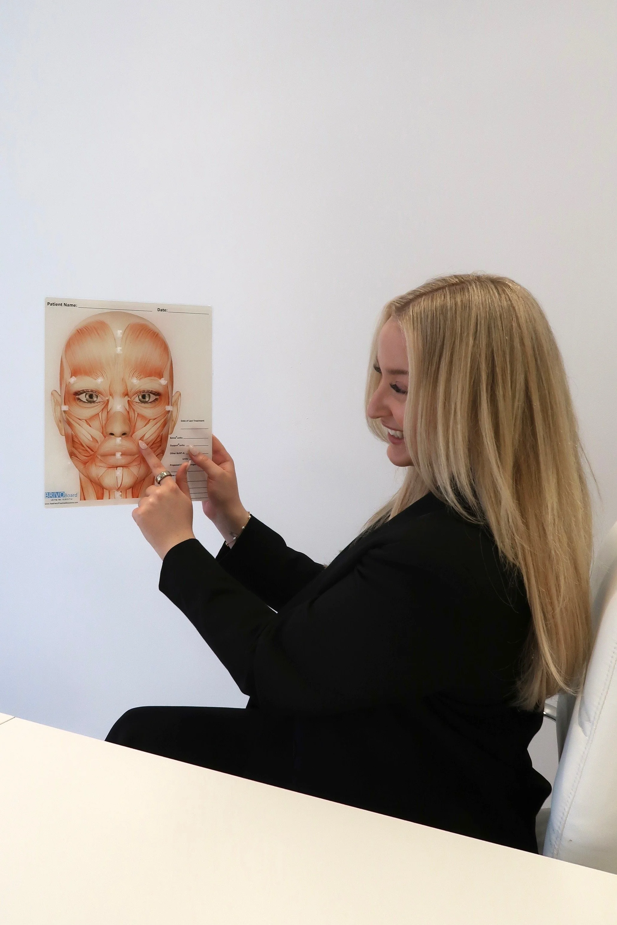 A woman with blonde hair holding a facial muscle diagram poster in a medical or educational setting.