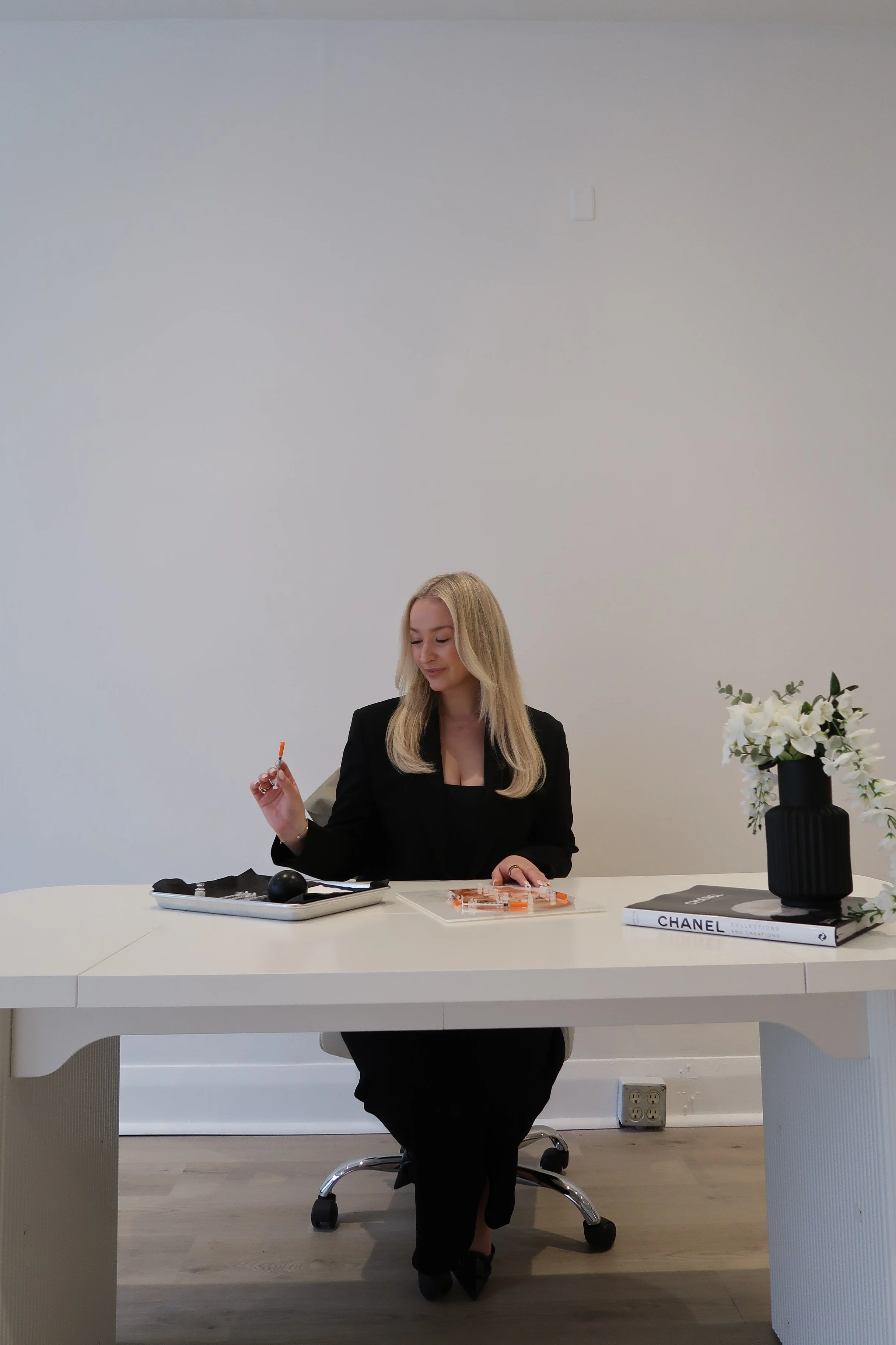 A woman with long blonde hair sits at a white table, holding an orange pen. She wears a black blazer and a low-cut top. On the table are a tray with stationery, a closed book labeled "CHANEL," and a vase with white flowers. The background is a plain 