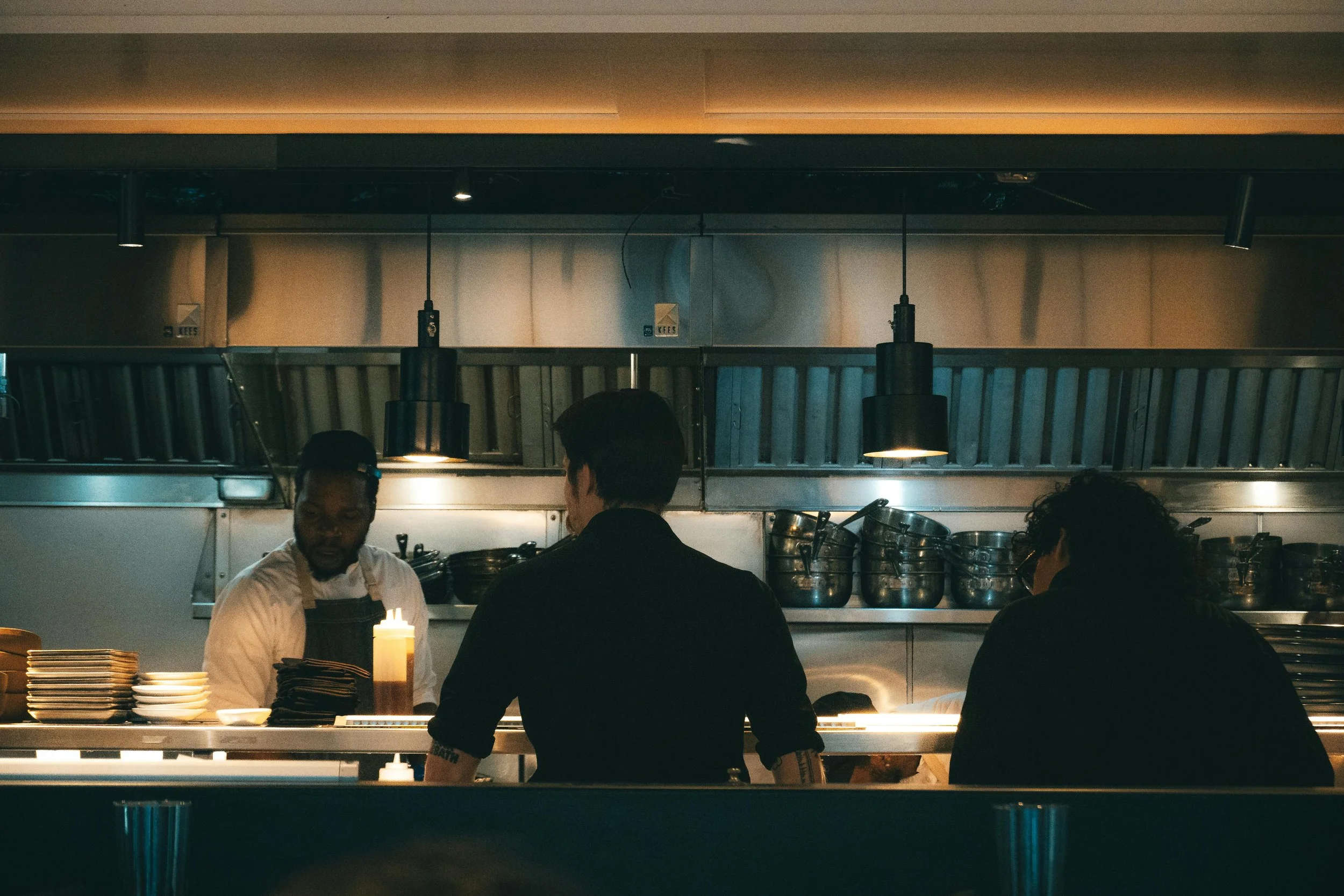 People working behind a restaurant counter, with plates and cooking utensils visible, in a kitchen with stainless steel appliances and hanging black pendant lights.