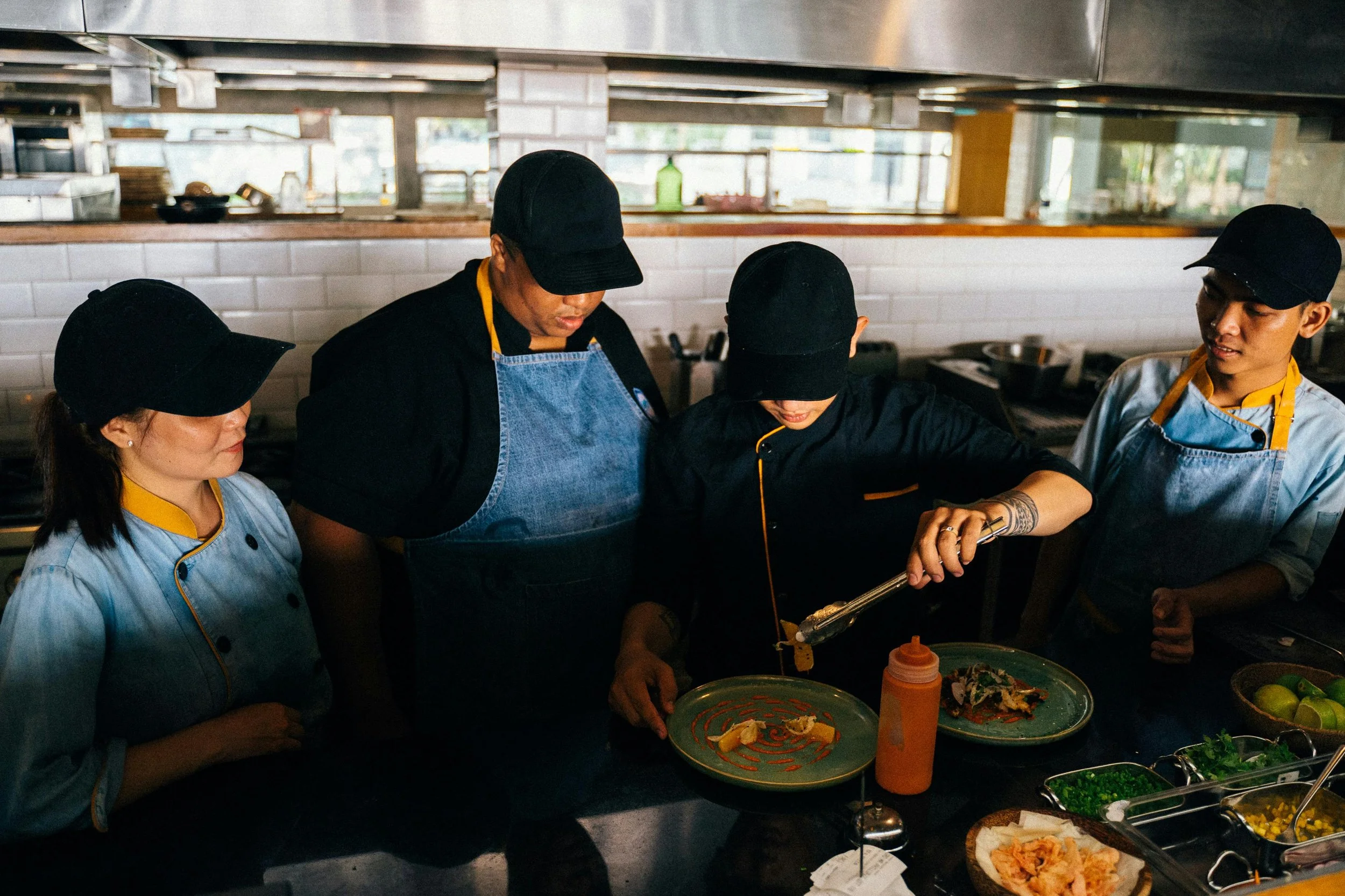 Four people working together in a restaurant kitchen, preparing or plating food, with various dishes and ingredients on the counter in front of them.