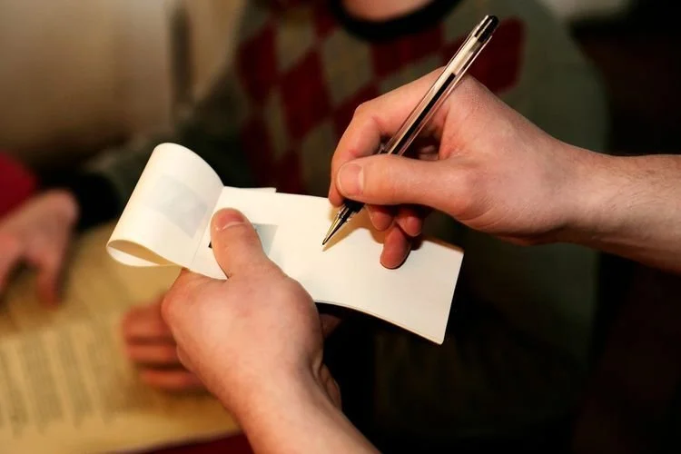 Person writing on a small spiral-bound notepad with a silver pen, another person is sitting nearby.