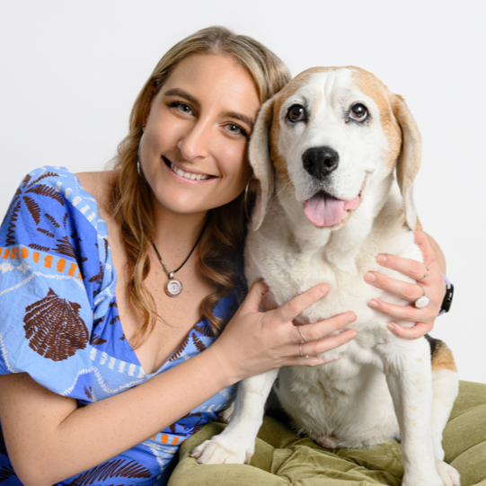 A woman with wavy blonde hair wearing a blue patterned top, smiling and hugging a large tan and white dog with blue eyes, sitting on a green cushion.