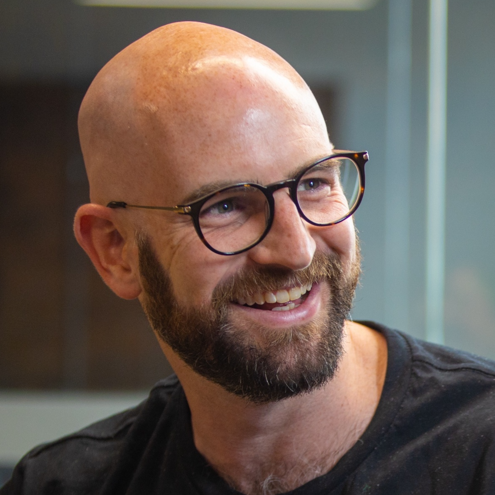 A smiling man with a beard and glasses, bald head, wearing a black shirt, looking to his right.
