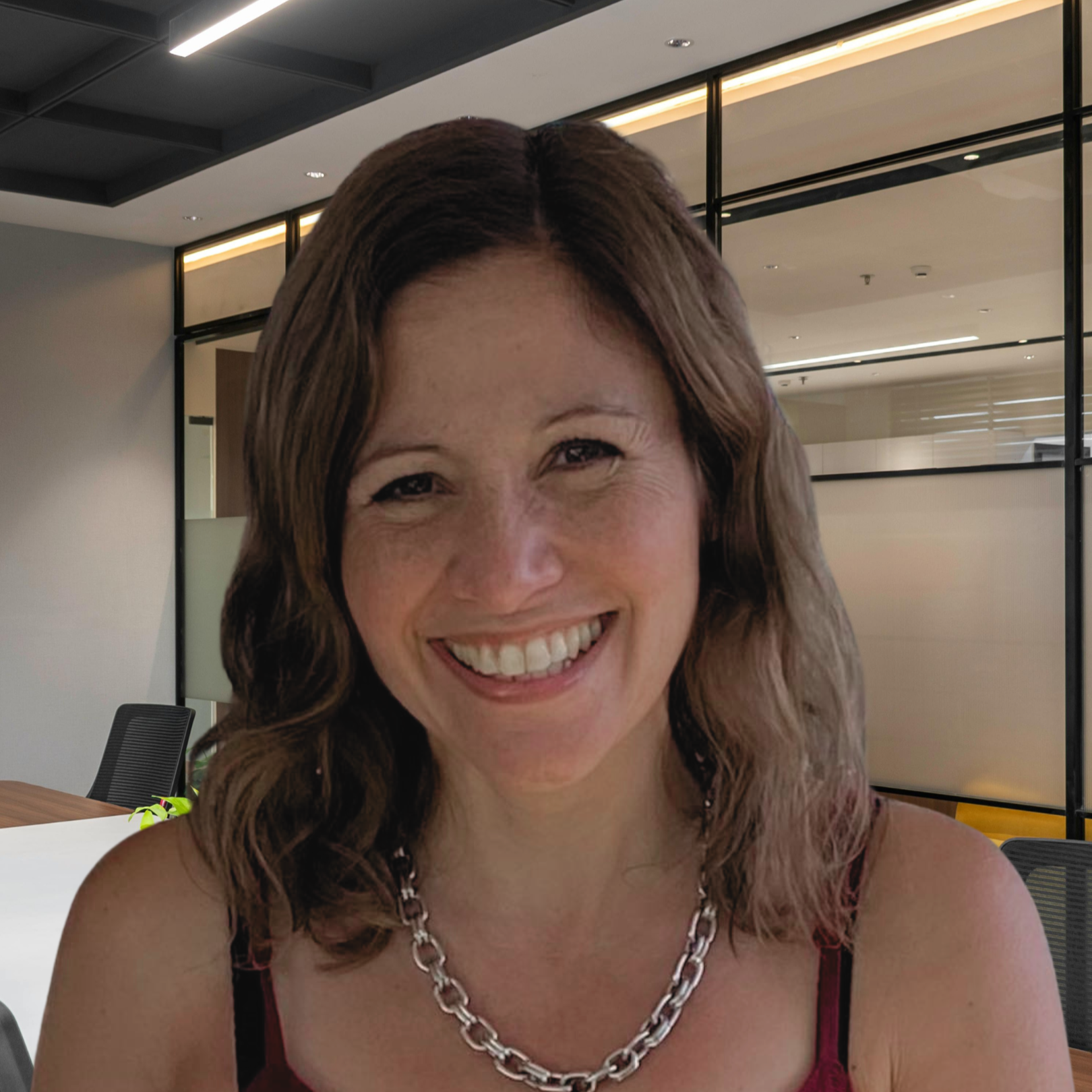 Close-up of a woman with wavy brown hair smiling, wearing a silver chain necklace and a red top, in a modern office setting.