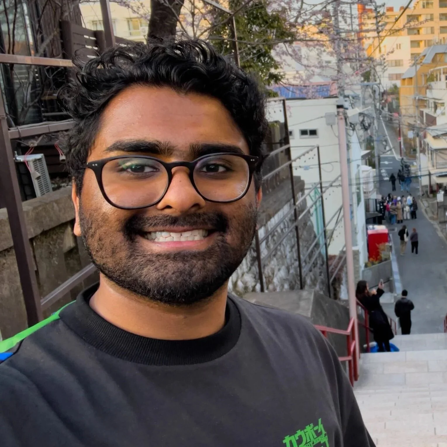 A smiling man with glasses and a beard taking a selfie outdoors on a staircase with an urban street scene in the background, including buildings, power lines, and people walking.
