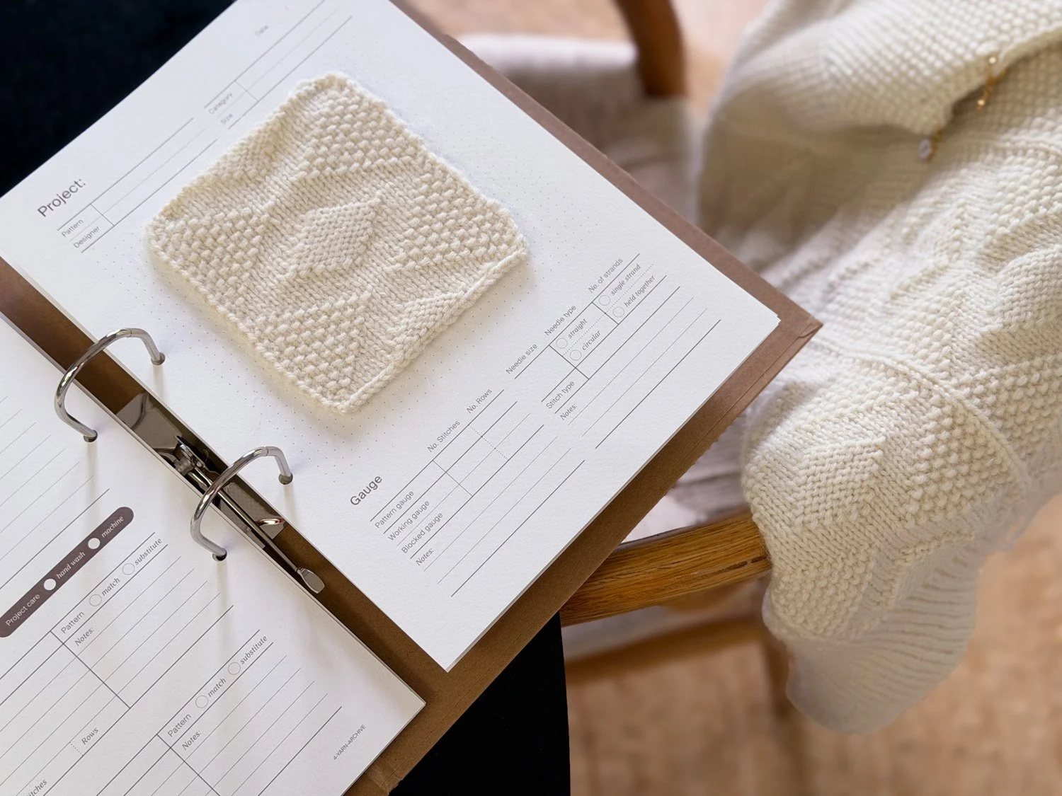 A knitting project worksheet with a swatch of cream-colored textured knitting and a partially completed knitted item resting on a wooden table.