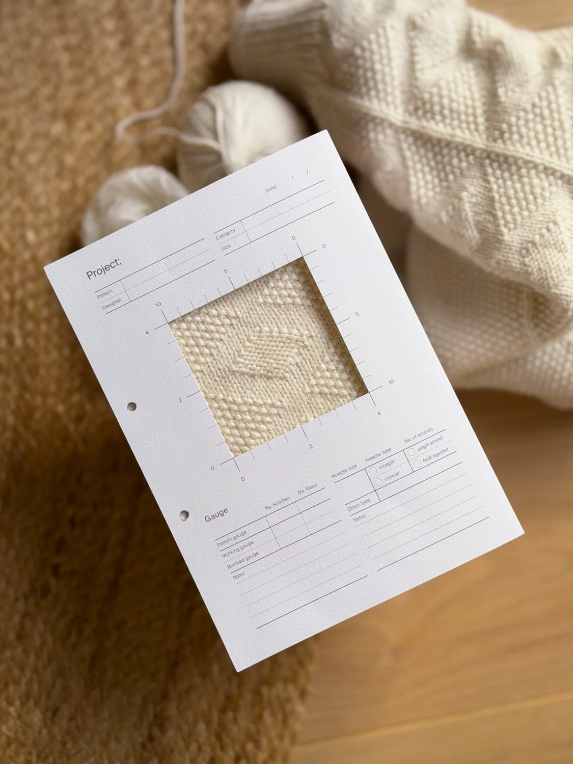 A knitting swatch inserted into a knitting project archive folder surrounded by knitting supplies, including partially finished knitted fabric and balls of yarn, on a wooden surface.
