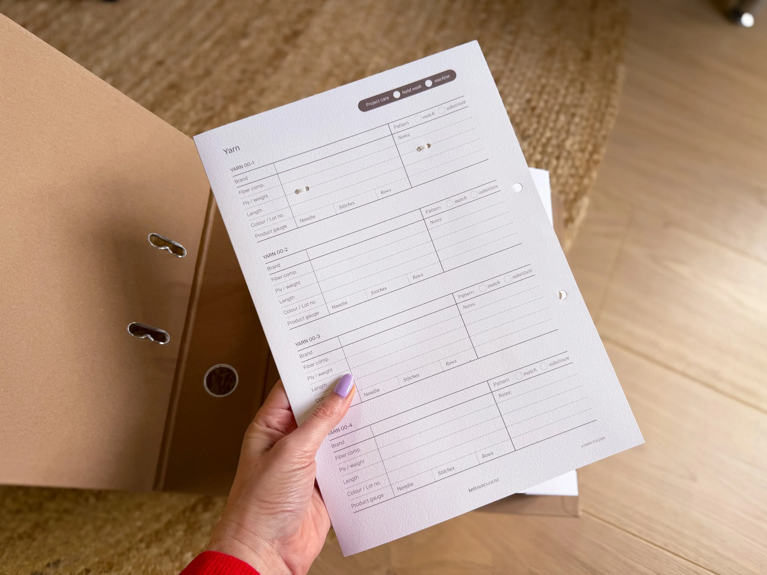 Person holding a printed form with knitting yarn detailed information, with a brown folder on a wooden surface in the background.