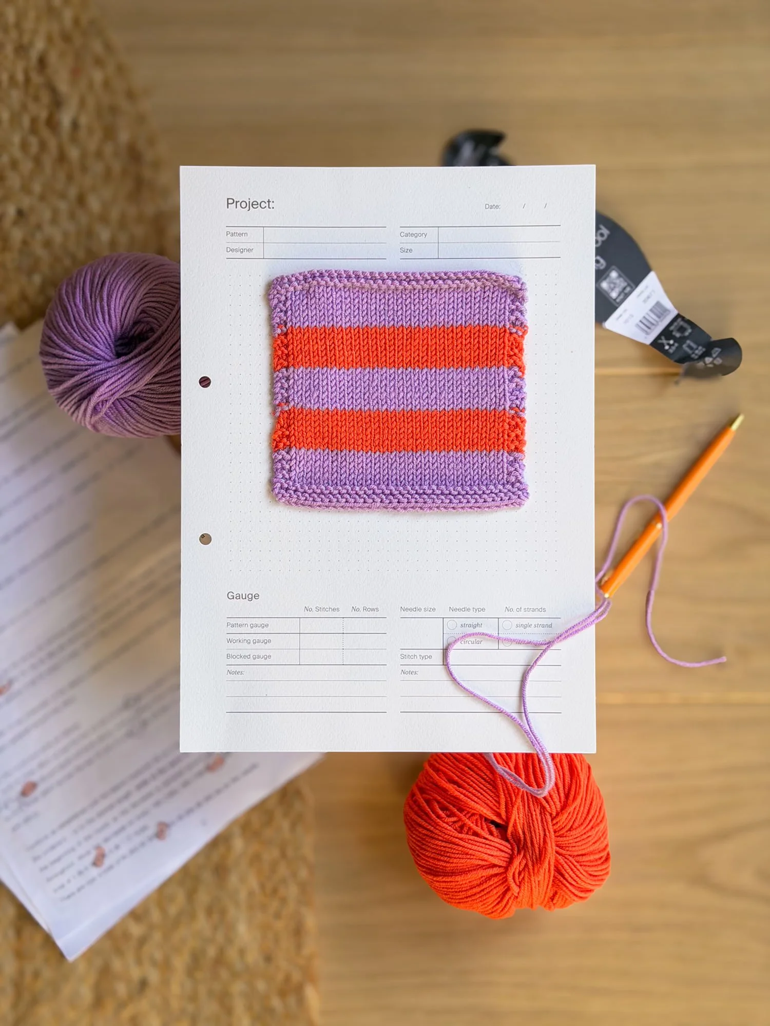 A knitting swatch with purple and orange stripes attached to a white project sheet with blank fields. A ball of orange yarn, a black yarn label, and a crochet hook are on a wooden surface.