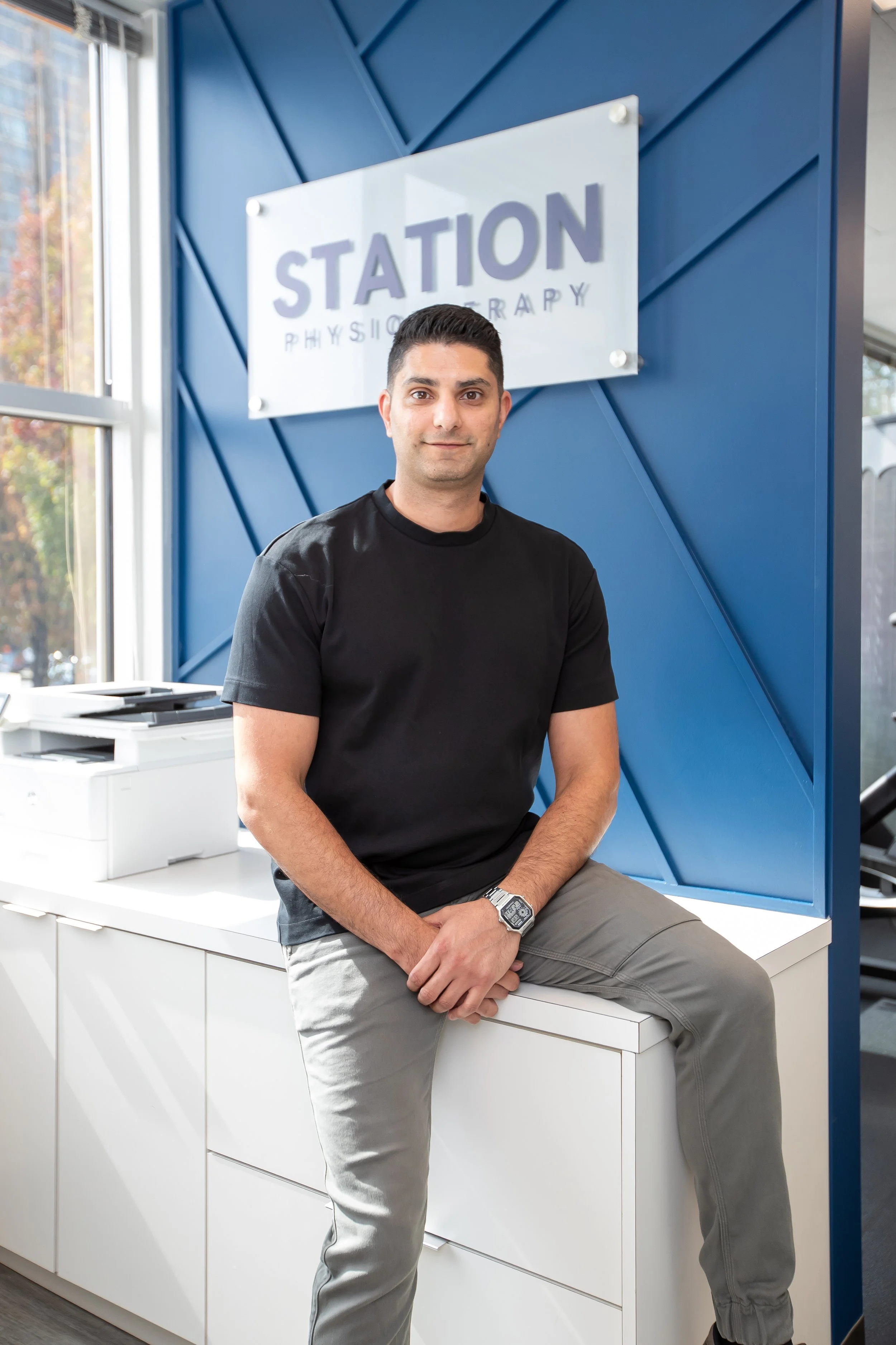 A man with dark hair and a casual black t-shirt sitting on a white cabinet at a therapy or rehab clinic, with a sign reading 'STATION PHYSIOTHERAPY' on a blue wall behind him.