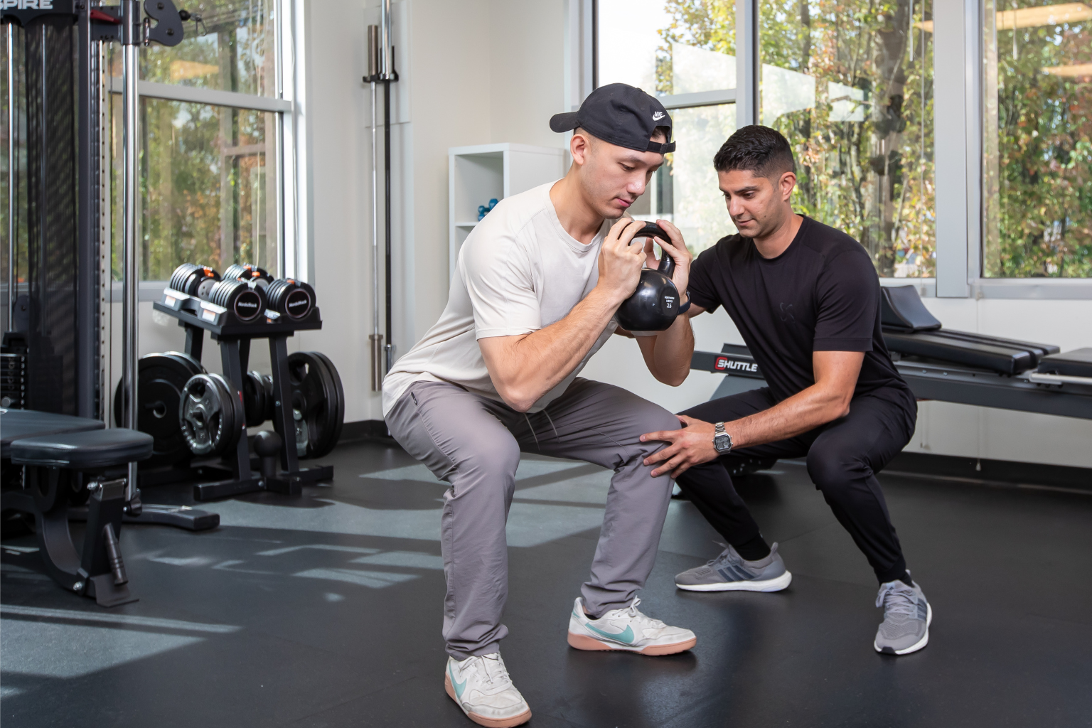 A personal trainer assisting a young man with a kettlebell squat exercise in a gym, with workout equipment and large windows in the background.