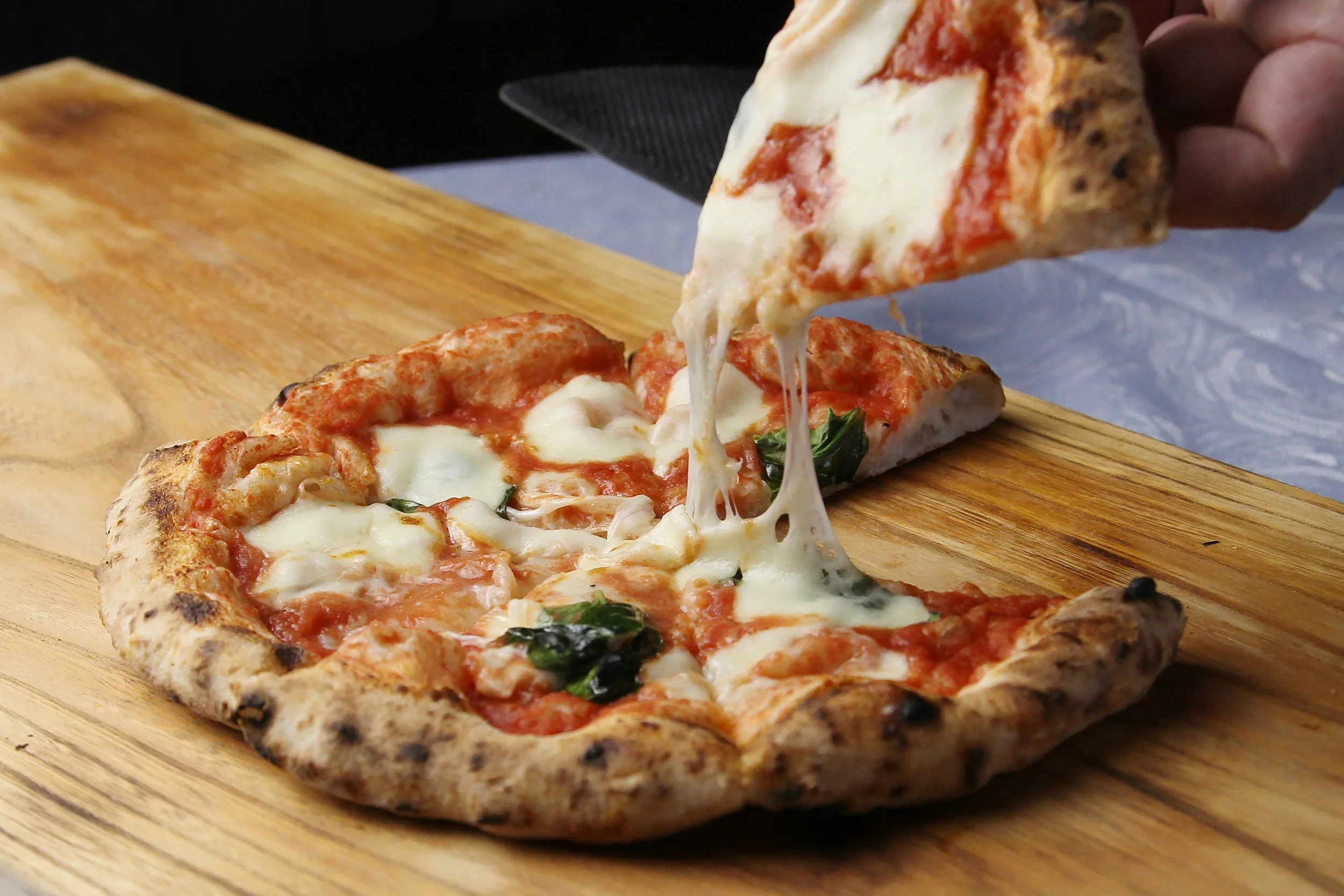 Close-up of a slice of cheese pizza with basil on a wooden board, with another slice being lifted by a hand, stretching melted cheese.